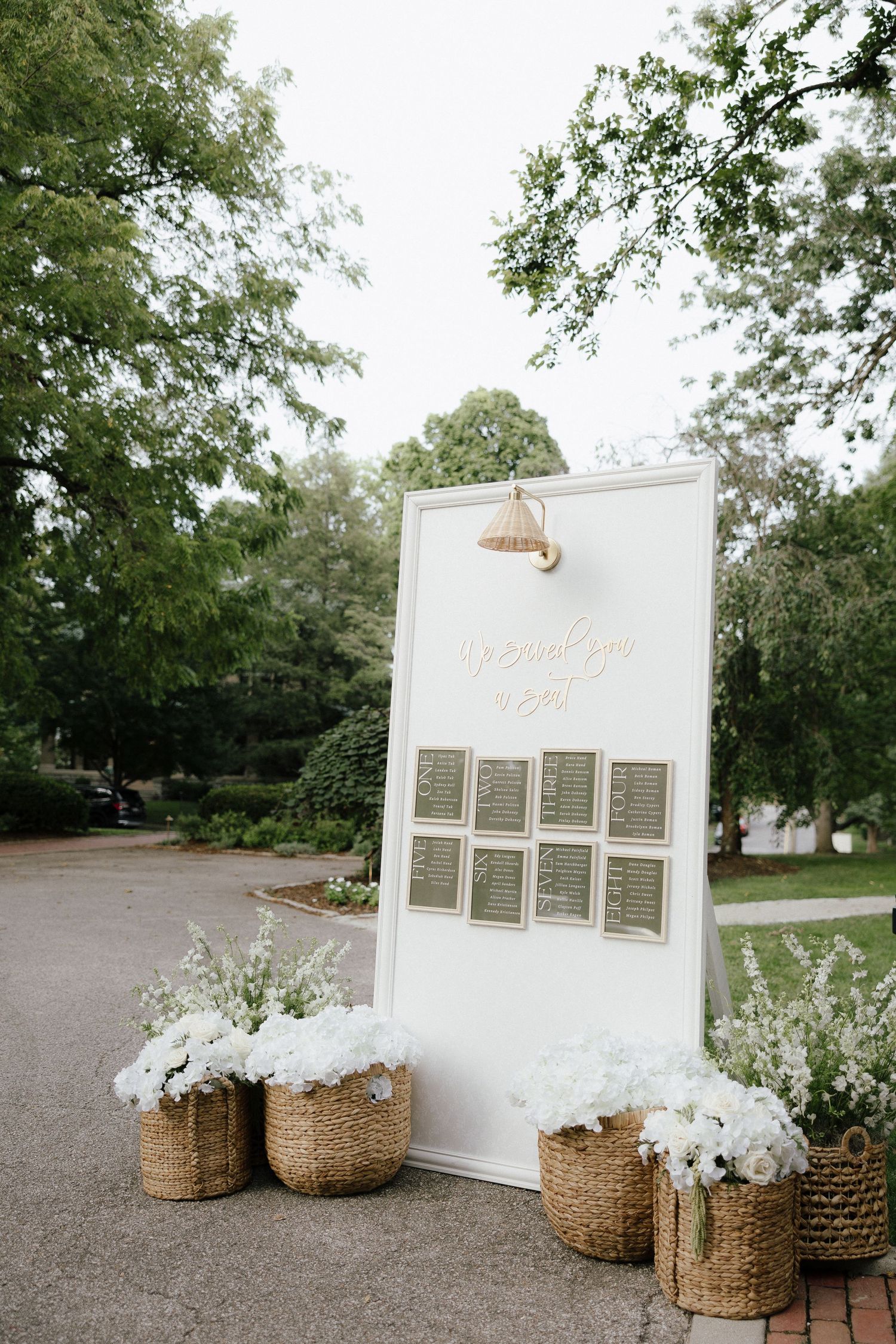 White wedding seating chart display board with wicker baskets of white flowers in an outdoor garden.