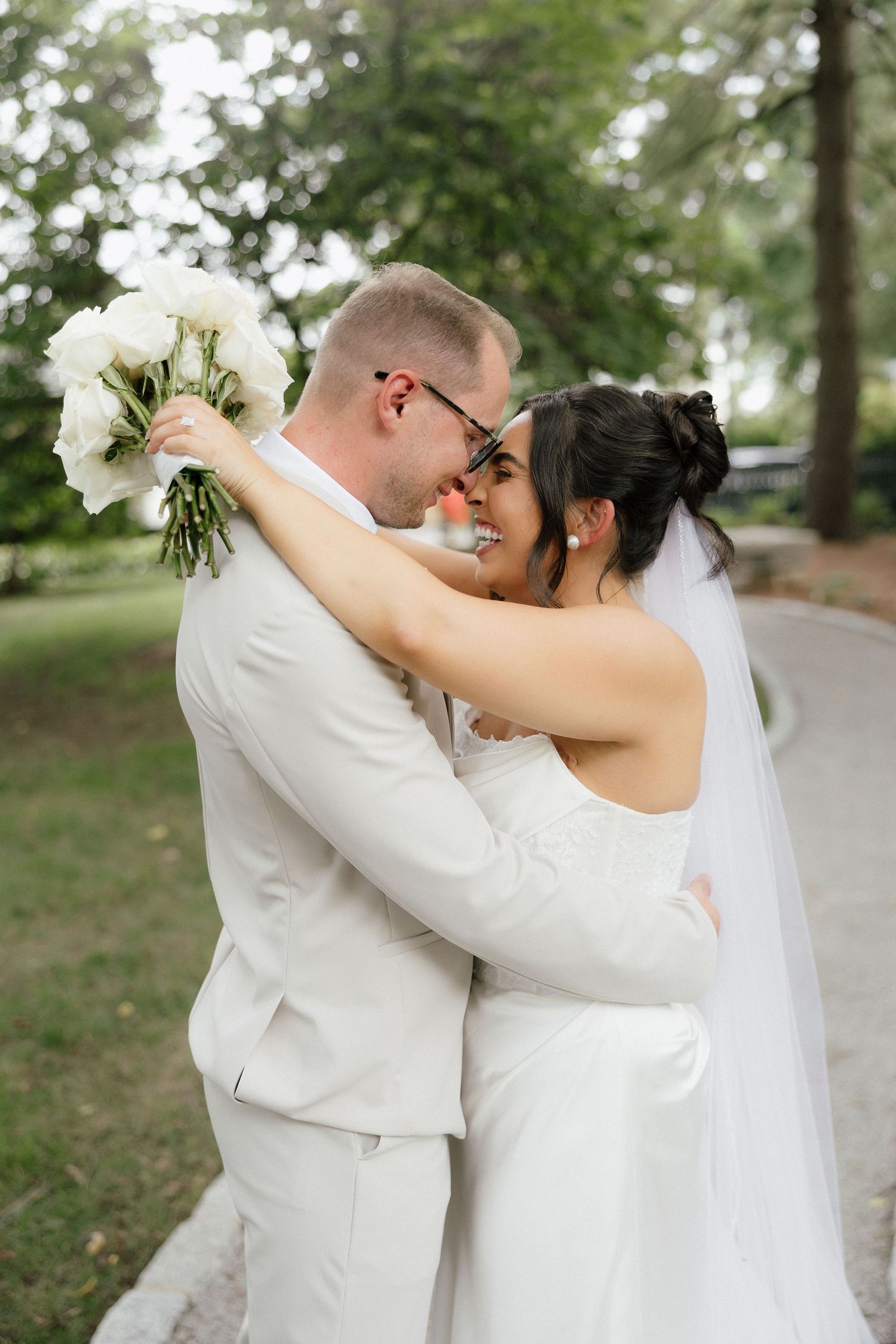 Bride and groom embrace and touch foreheads outdoors, bride holds white bouquet in elegant wedding attire.