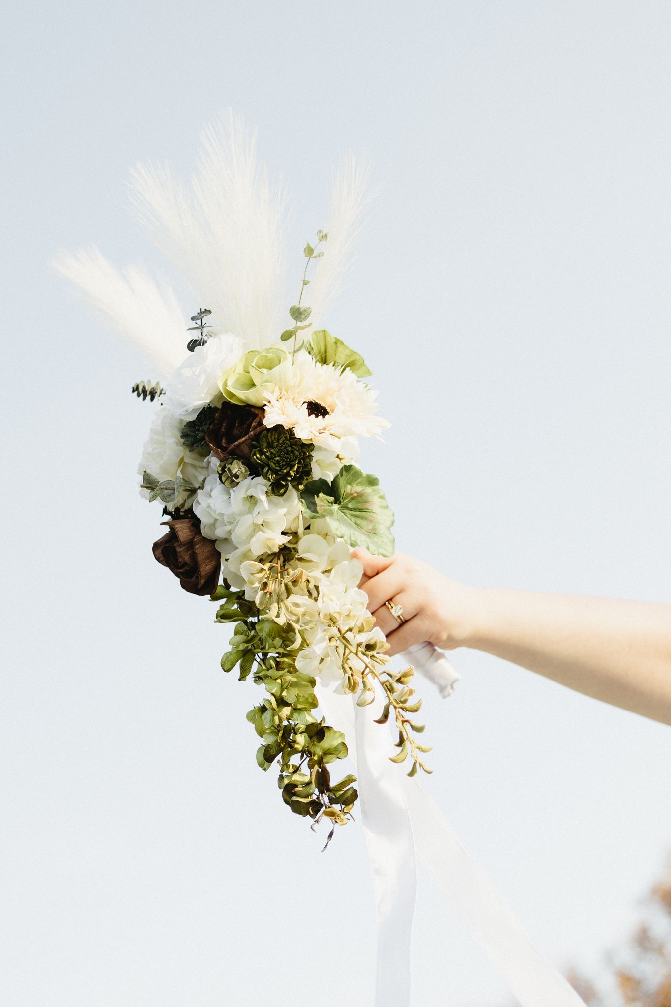 A hand holds a delicate white floral bouquet against a bright sky while bees hover around the blooms.