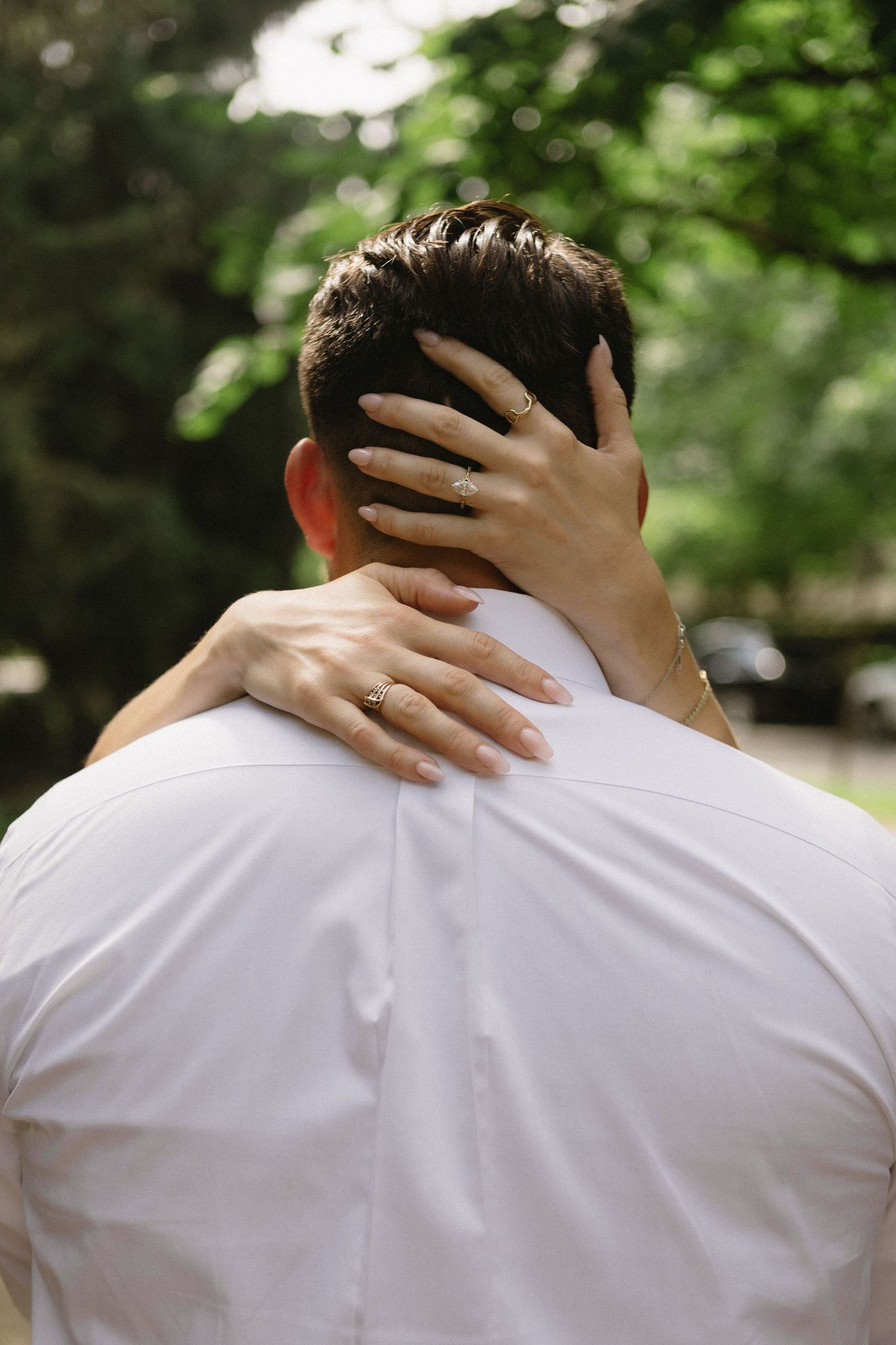 Close-up of bride's hands with engagement ring embracing groom's neck from behind during outdoor photo session.