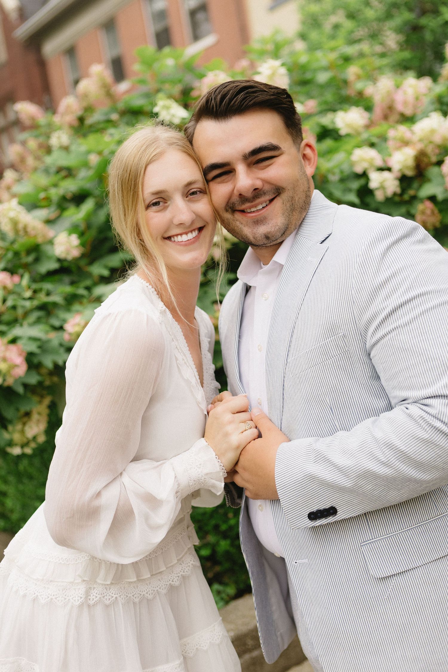 Smiling couple in white dress and gray suit embracing in front of blooming hydrangea bushes outdoors.