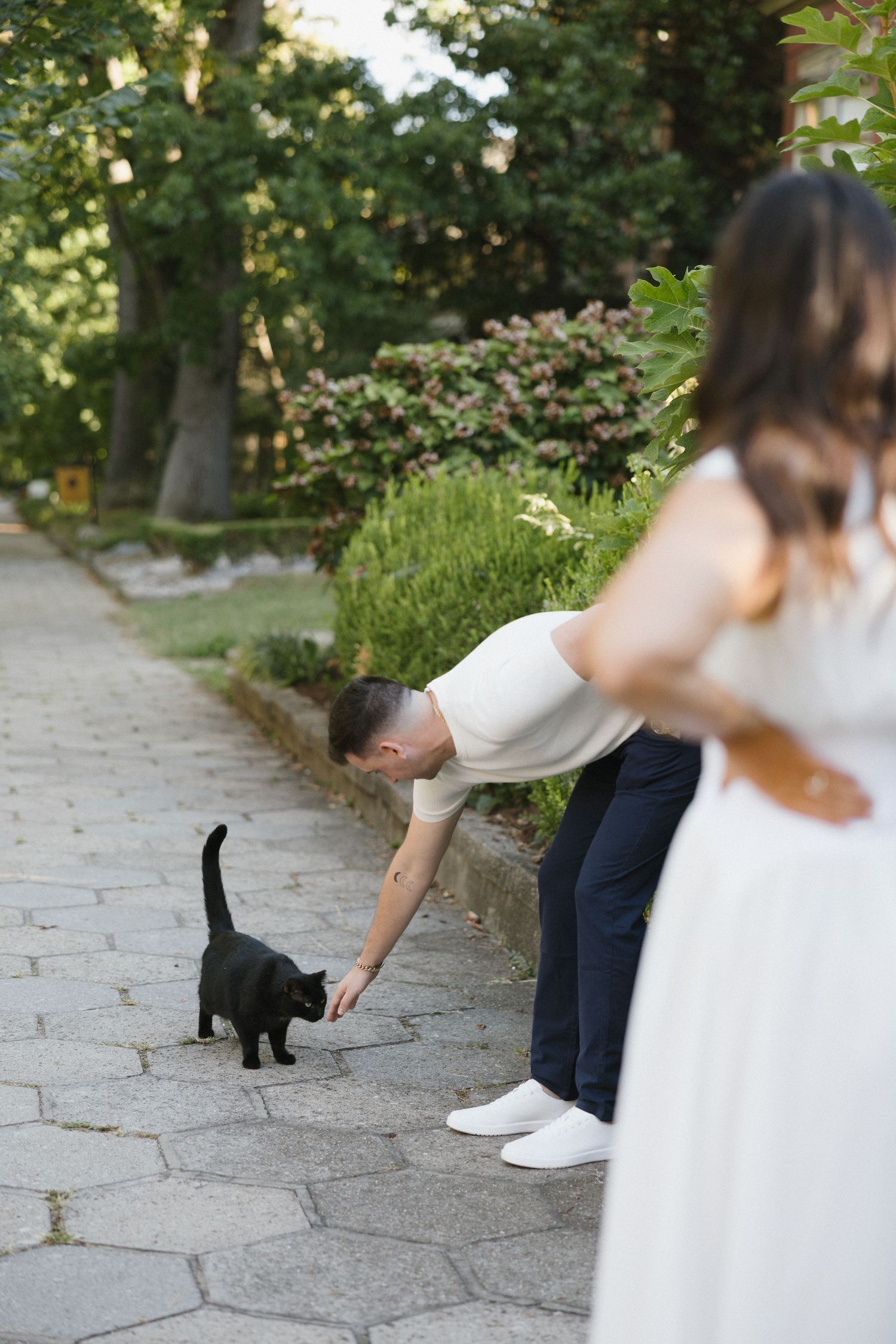 A man in white shirt bends to pet a black cat on a garden path while a woman in white dress stands nearby.