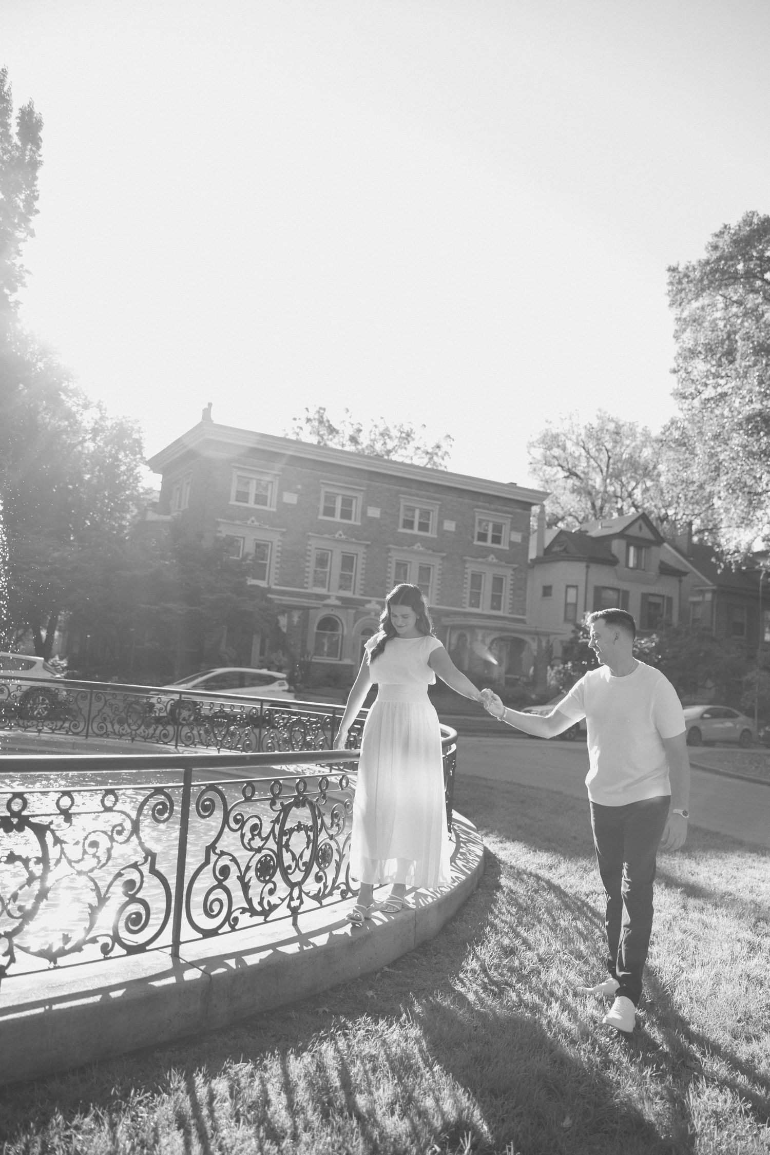 Black and white photo of a couple holding hands walking along an ornate iron fence in a sunny park.
