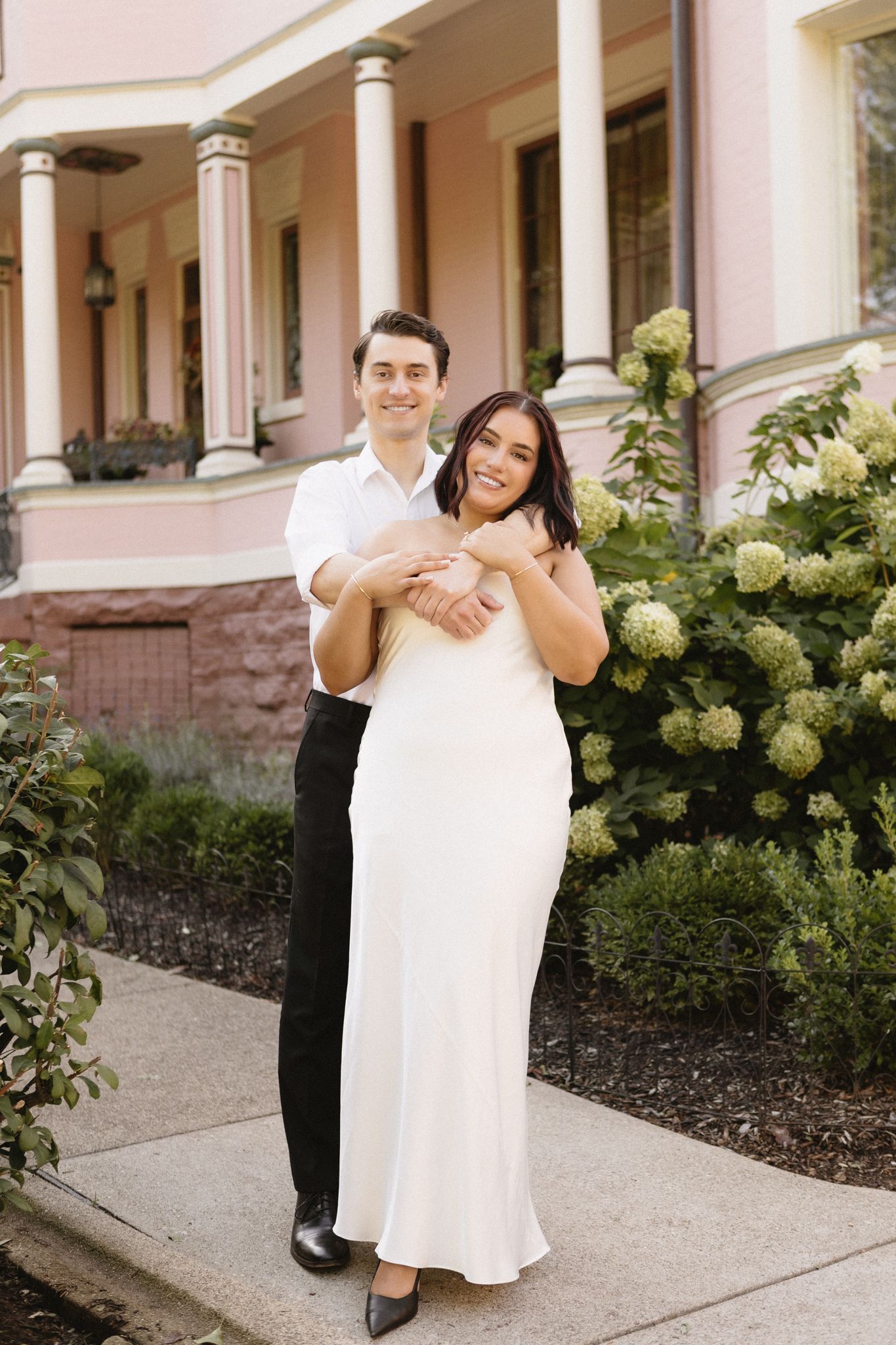 A romantic moment between a couple on a porch with pink columns and hydrangea bushes.