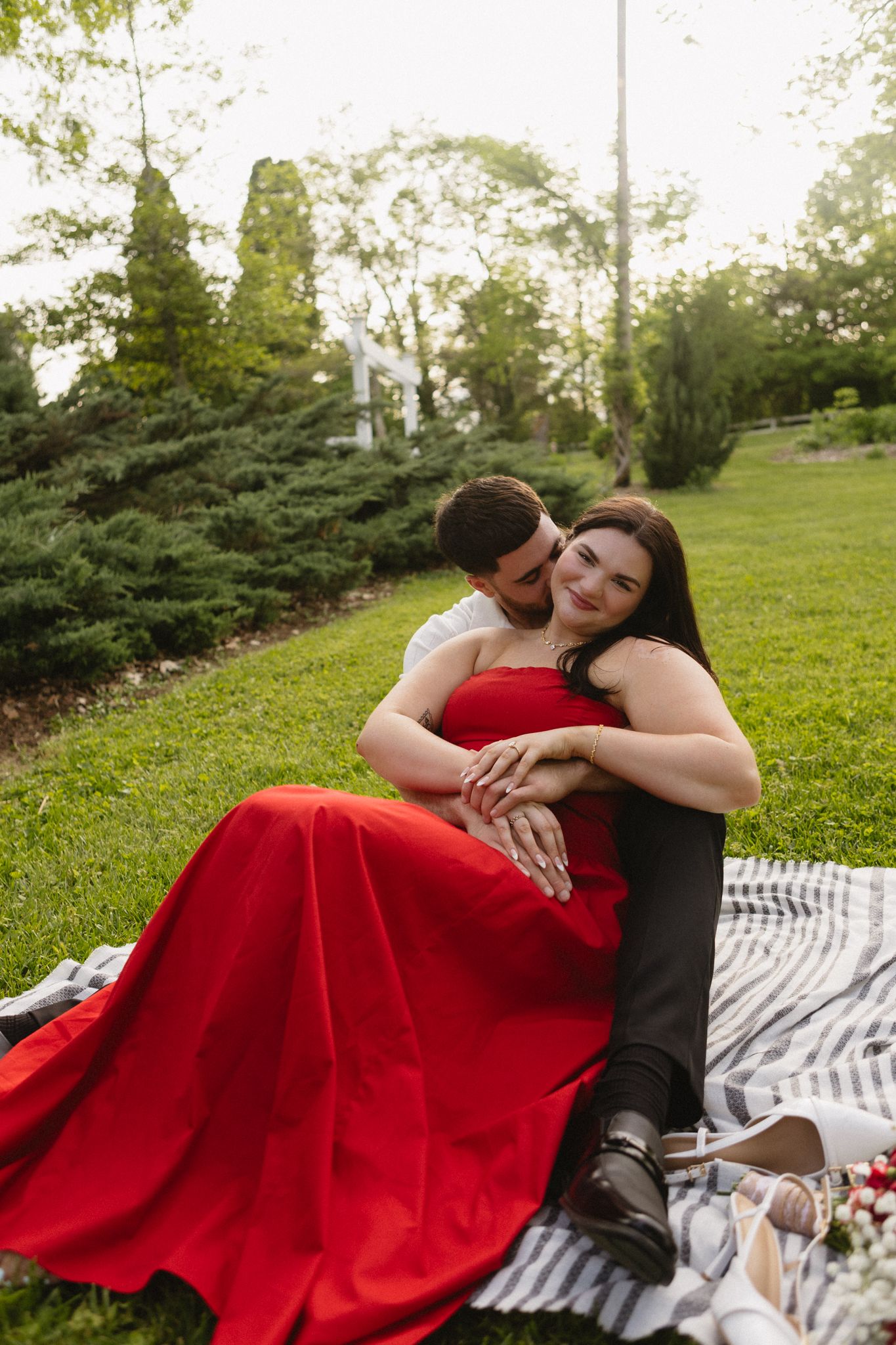 A couple in red and black attire share an intimate moment while sitting on a striped blanket in a grassy park setting.