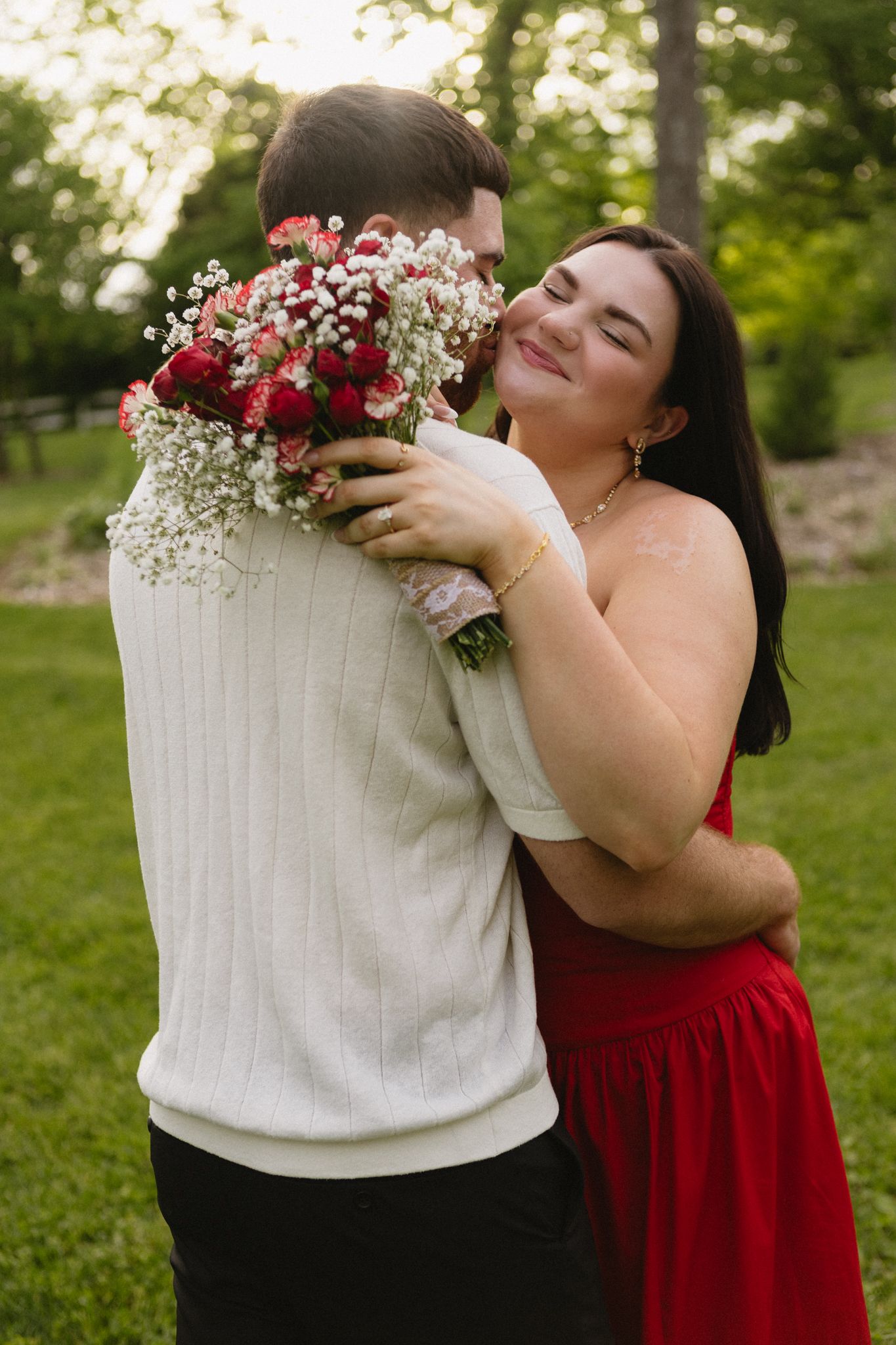 Two people share an embrace outdoors, with one wearing a vibrant red dress and holding a bouquet of mixed flowers.
