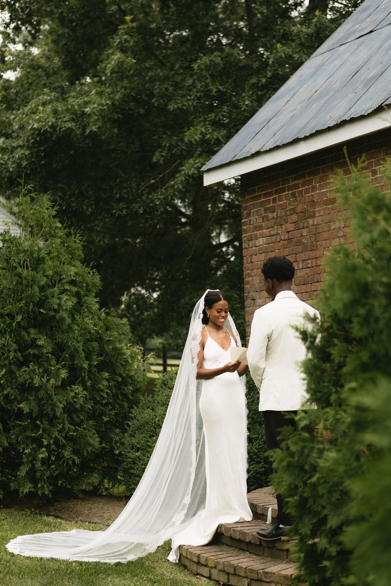 Wedding couple shares a tender moment outside a rustic brick building surrounded by greenery.