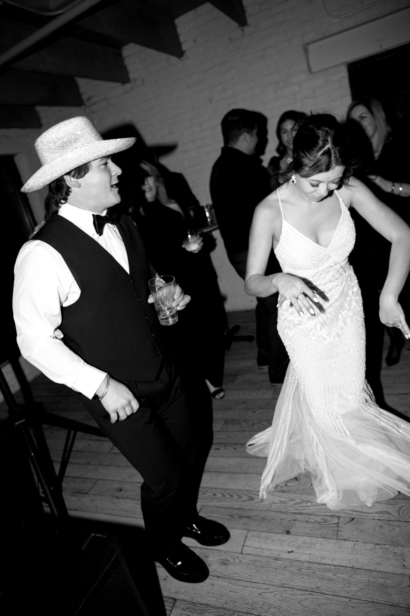 Wedding guests dance at a reception with one wearing a cowboy hat and the bride in a flowing white dress.