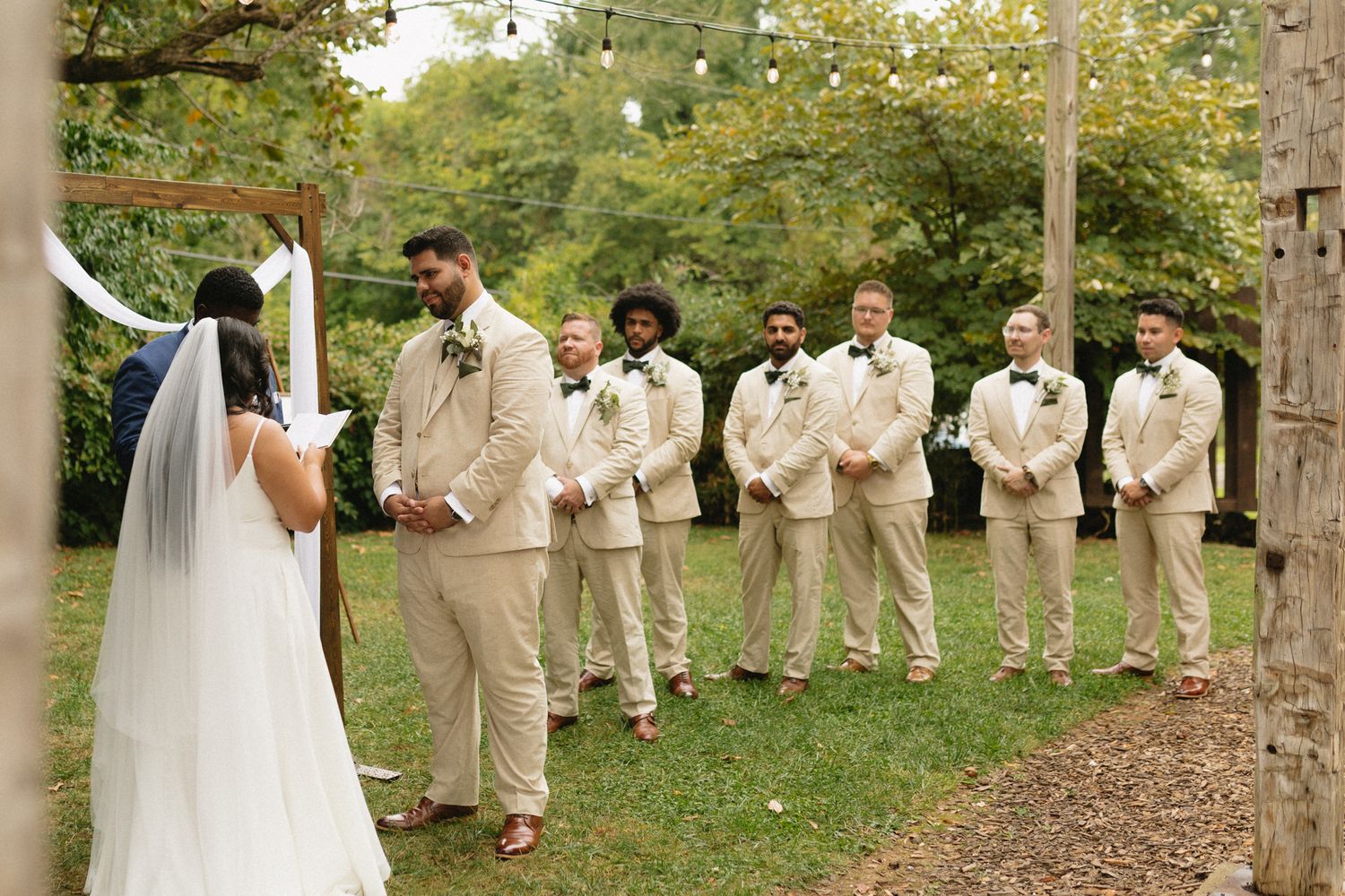 A wedding party stands outdoors in cream-colored suits while the bride in white faces them during a forest ceremony.