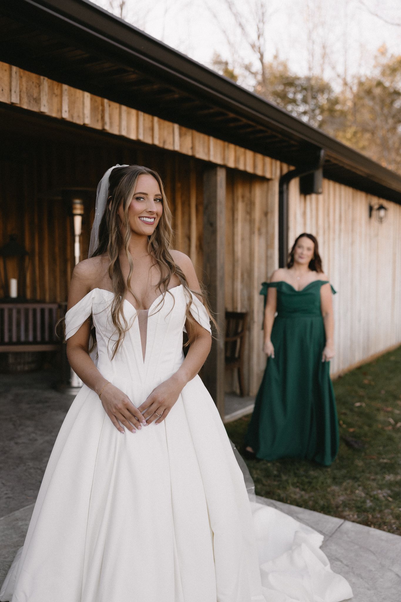 Elegant white wedding dress with flowing train and bridesmaids in emerald green dresses in rustic barn setting.