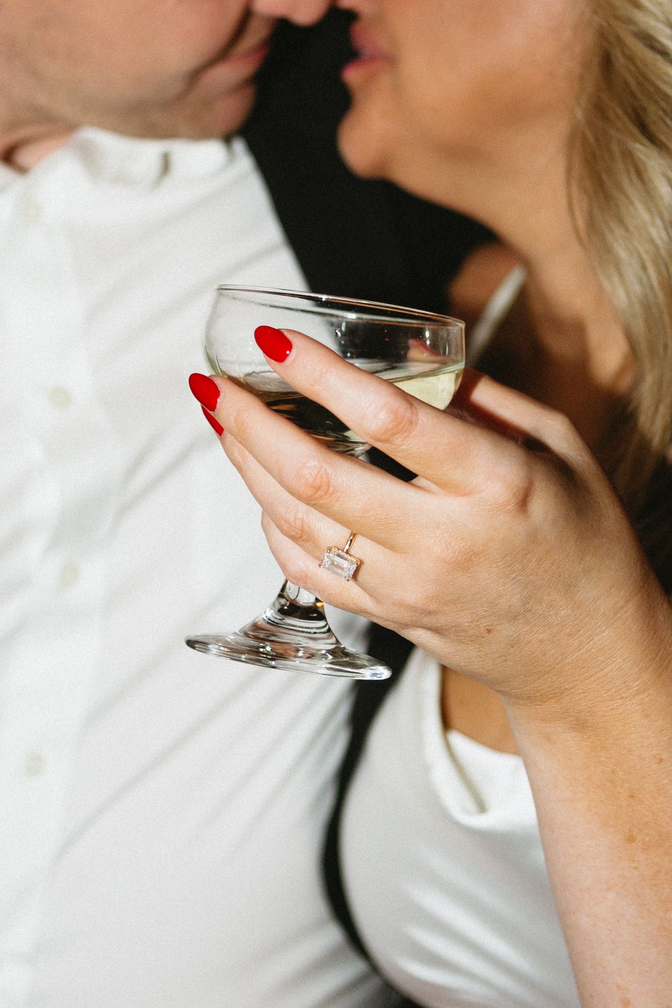 Close-up of hands holding champagne glass with red nails and engagement ring during romantic moment.