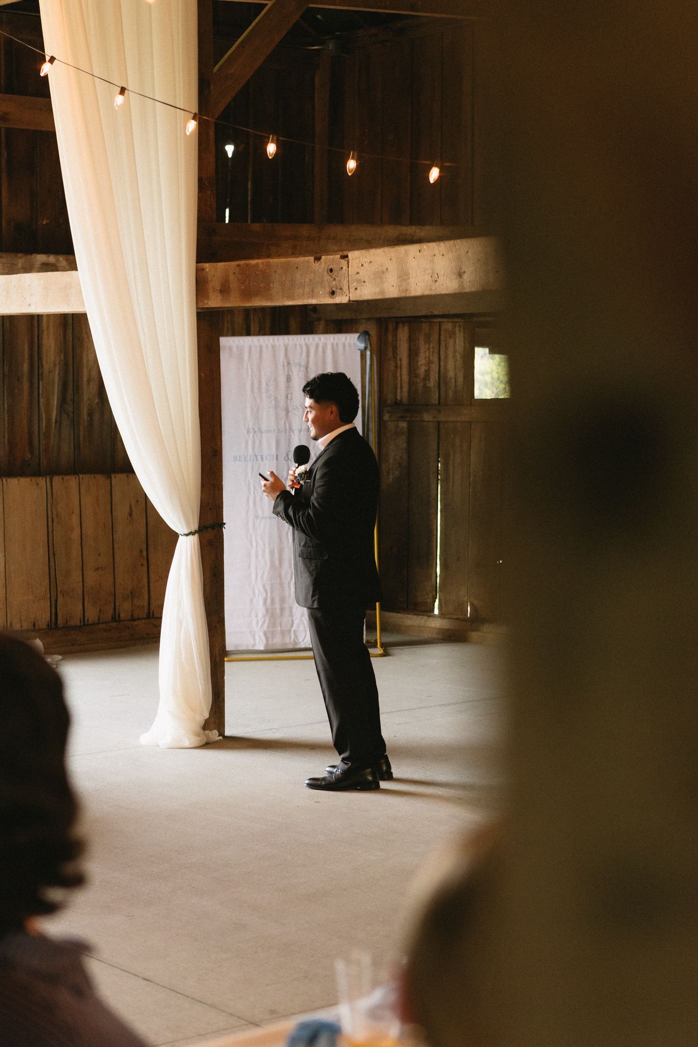 Speaker delivers wedding speech in dimly lit barn venue with white curtain backdrop.