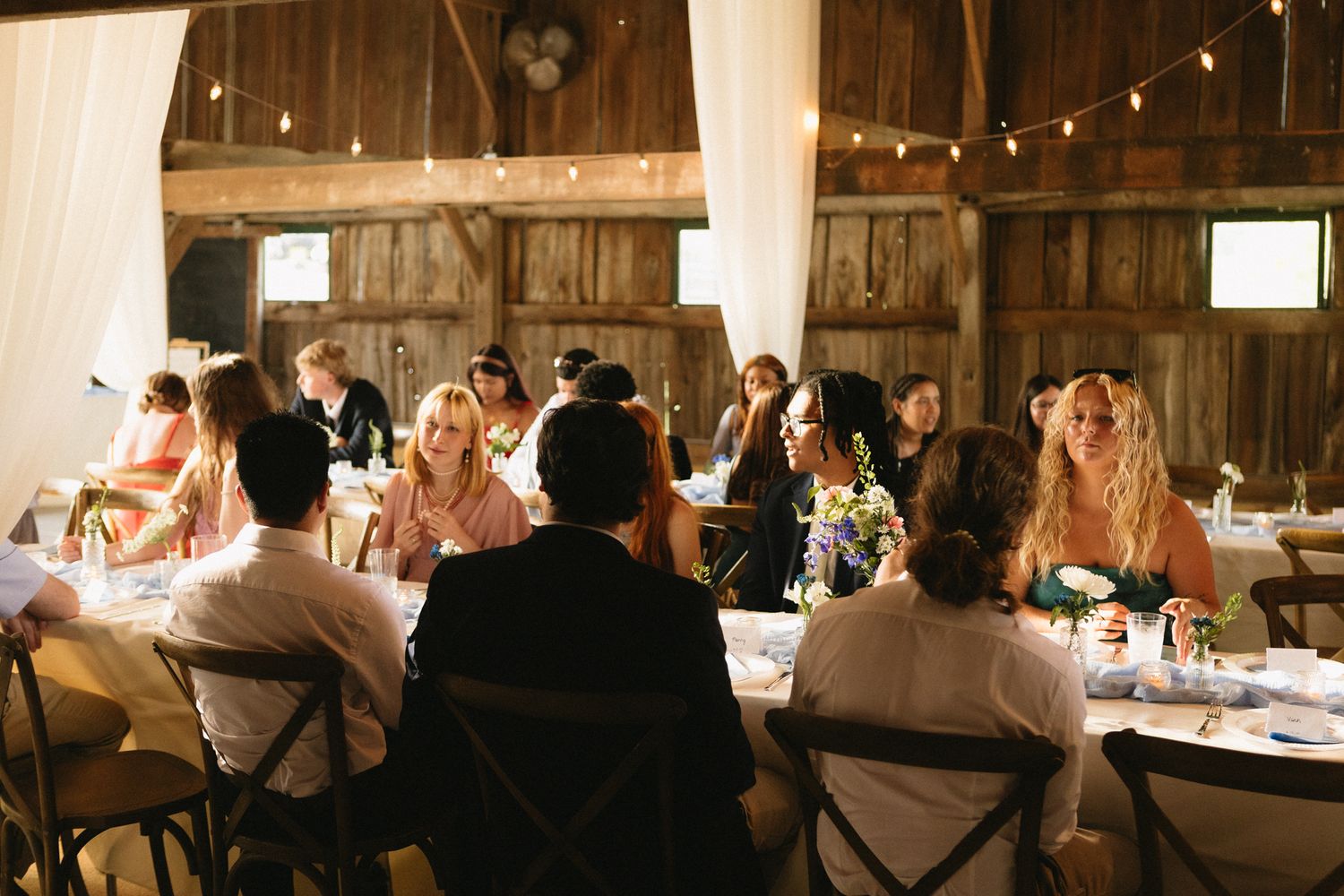 Reception dinner scene inside rustic barn venue with string lights and wooden beams.