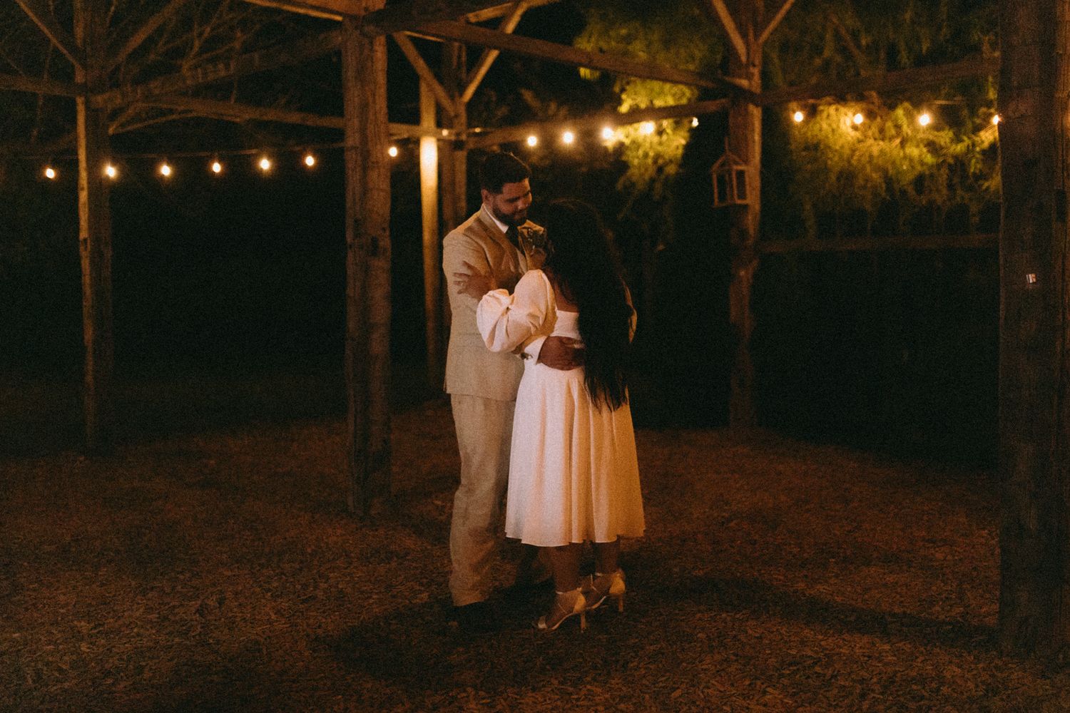 A romantic evening embrace under warm string lights in a rustic outdoor setting.