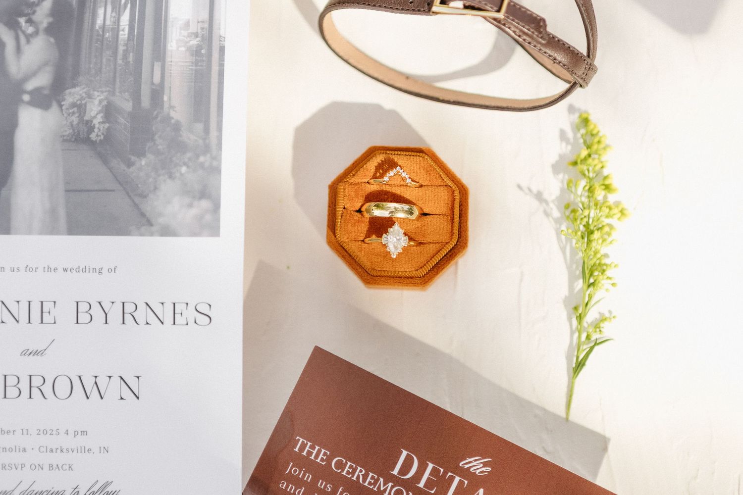 Wooden octagonal ring box displayed with wedding invitation and delicate green floral sprig on white surface.