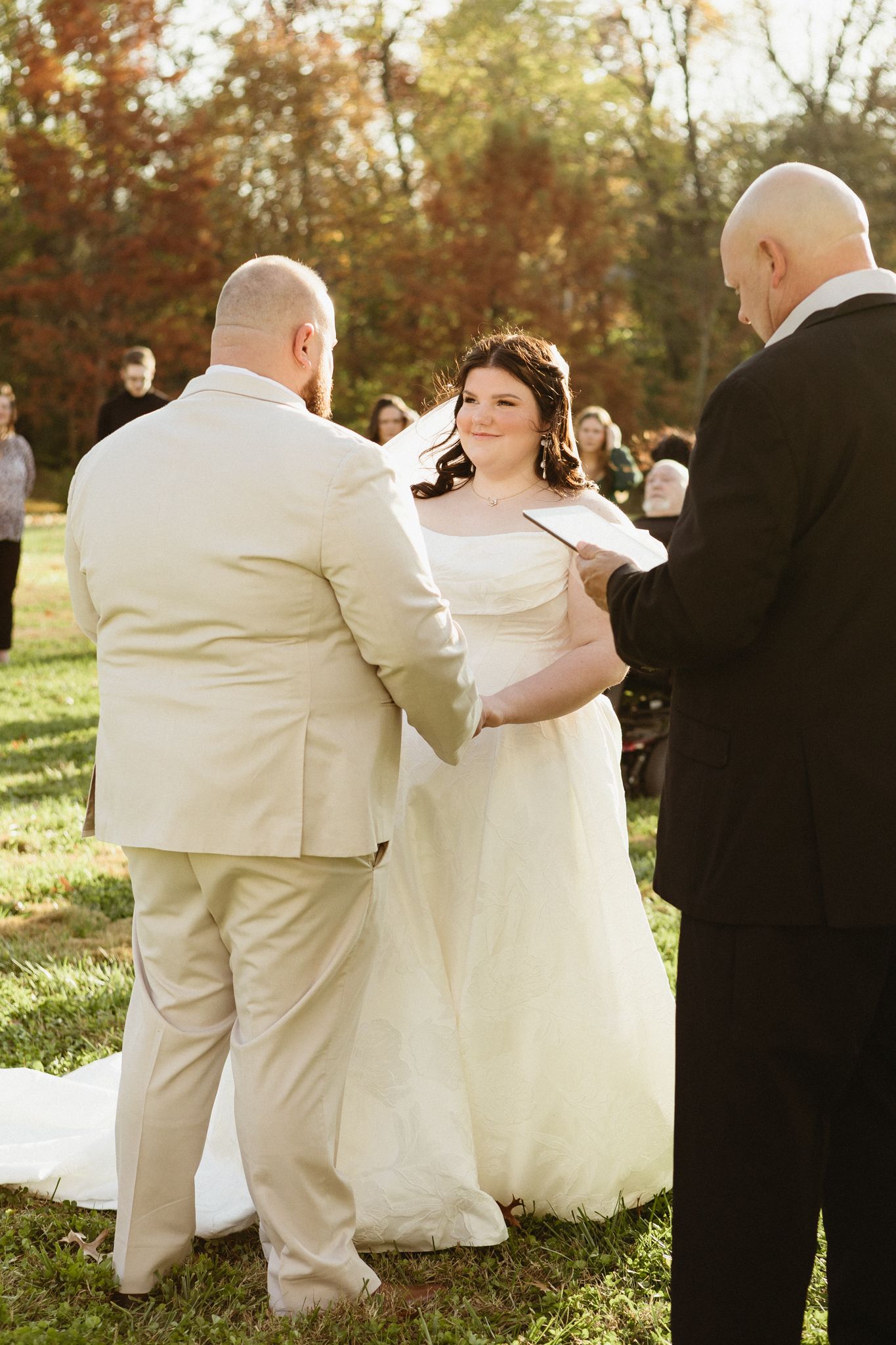 Wedding party members in light colored suits and dark dresses pose together during golden hour at an outdoor ceremony.
