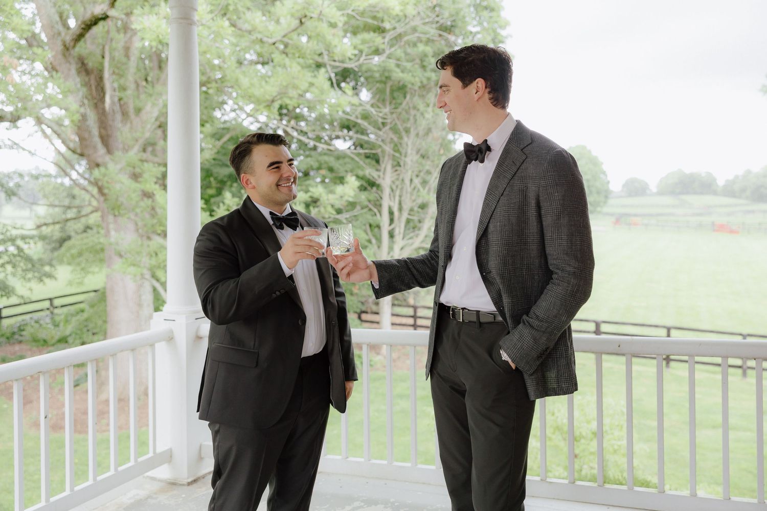 Two men in formal suits shaking hands on a white porch at an outdoor wedding venue.