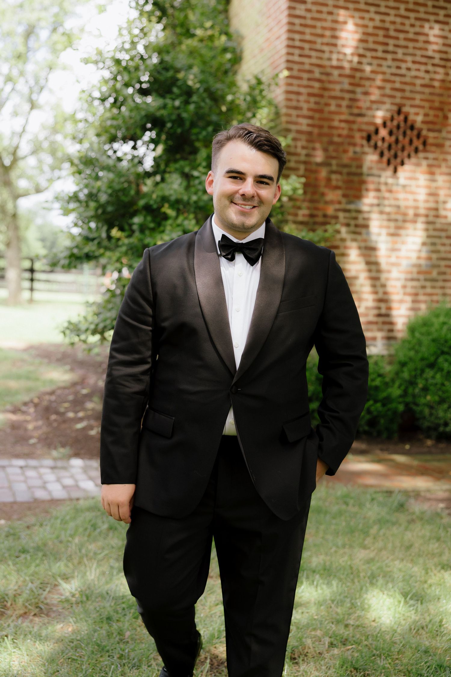 Smiling groom in black tuxedo with bow tie poses outdoors near brick wall and green trees.