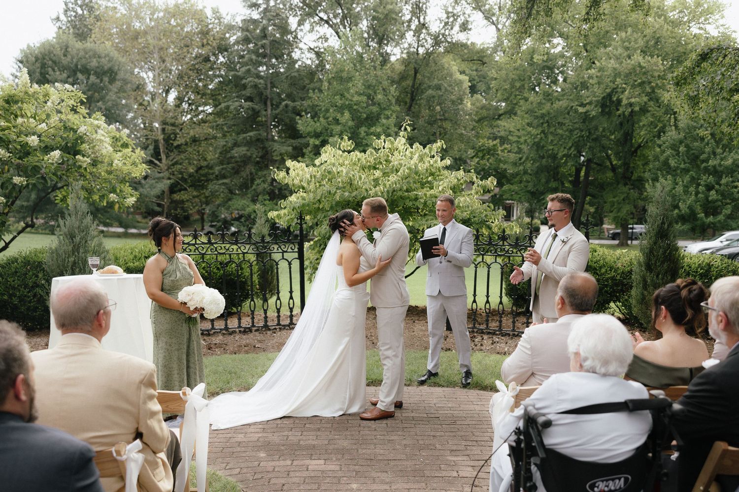 Bride and groom share first kiss at outdoor garden ceremony surrounded by guests and lush greenery.