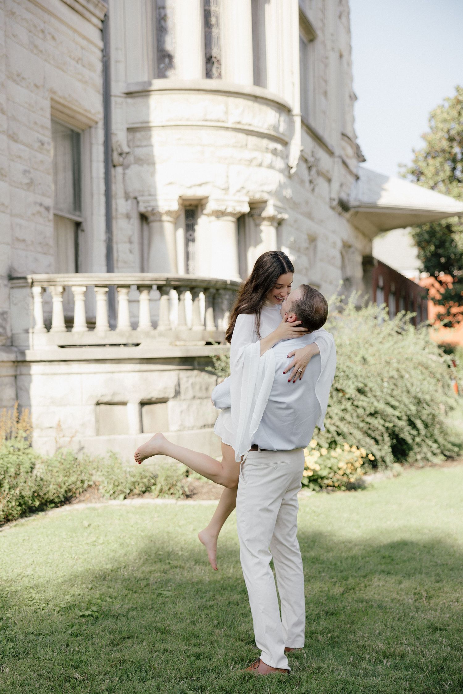 Romantic couple embracing and kissing in front of an elegant stone mansion on a sunny day.
