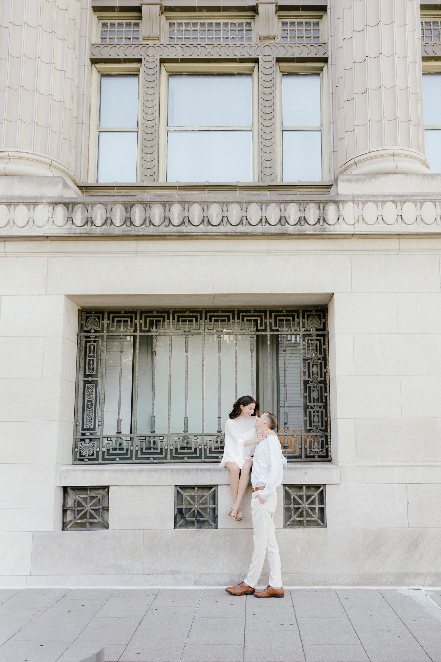 Couple sharing a tender kiss against an ornate beige stone building facade on a sunny day.