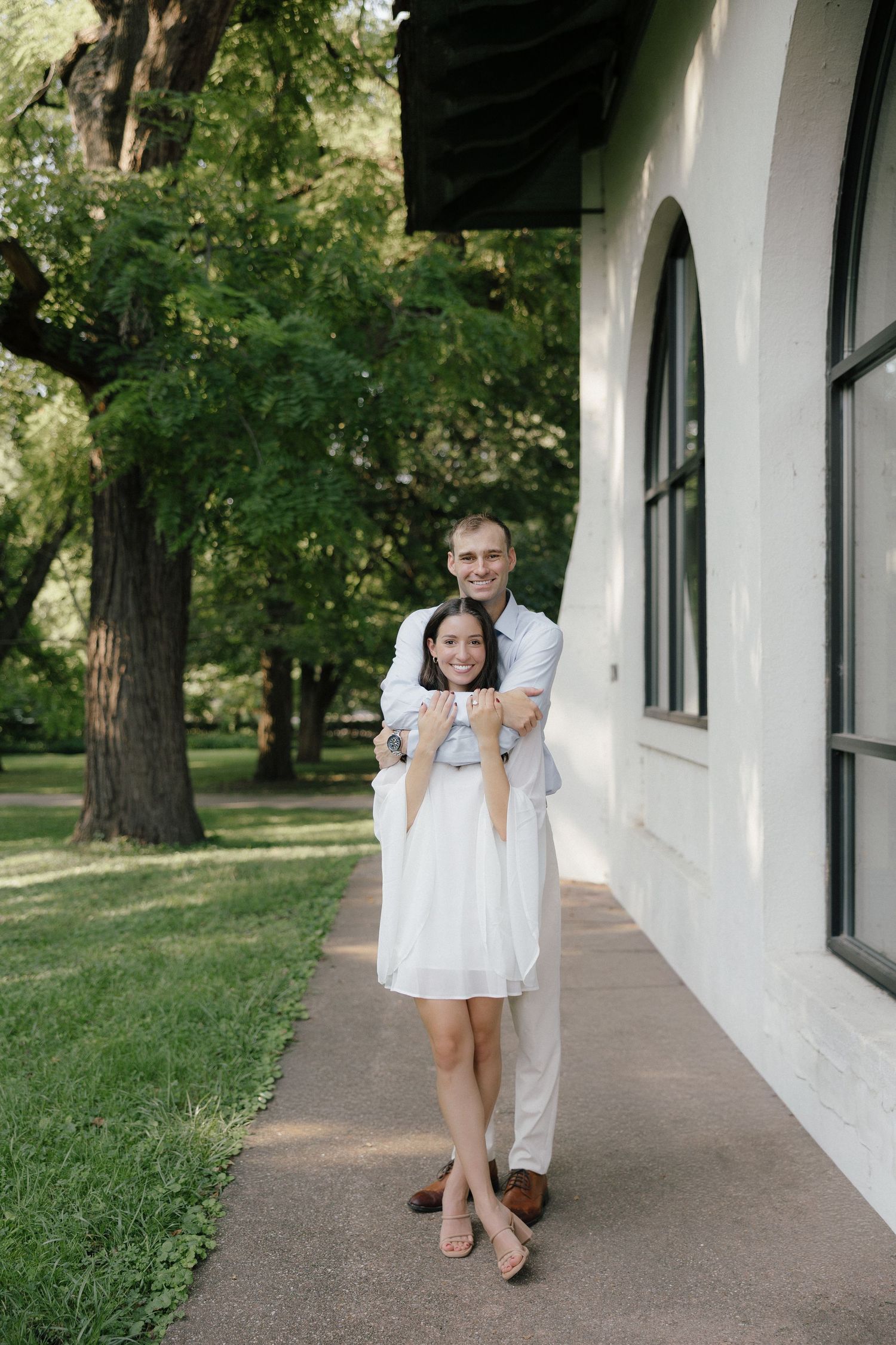 Happy couple embracing outdoors near white building, man hugging woman in white dress, lush green trees in background.