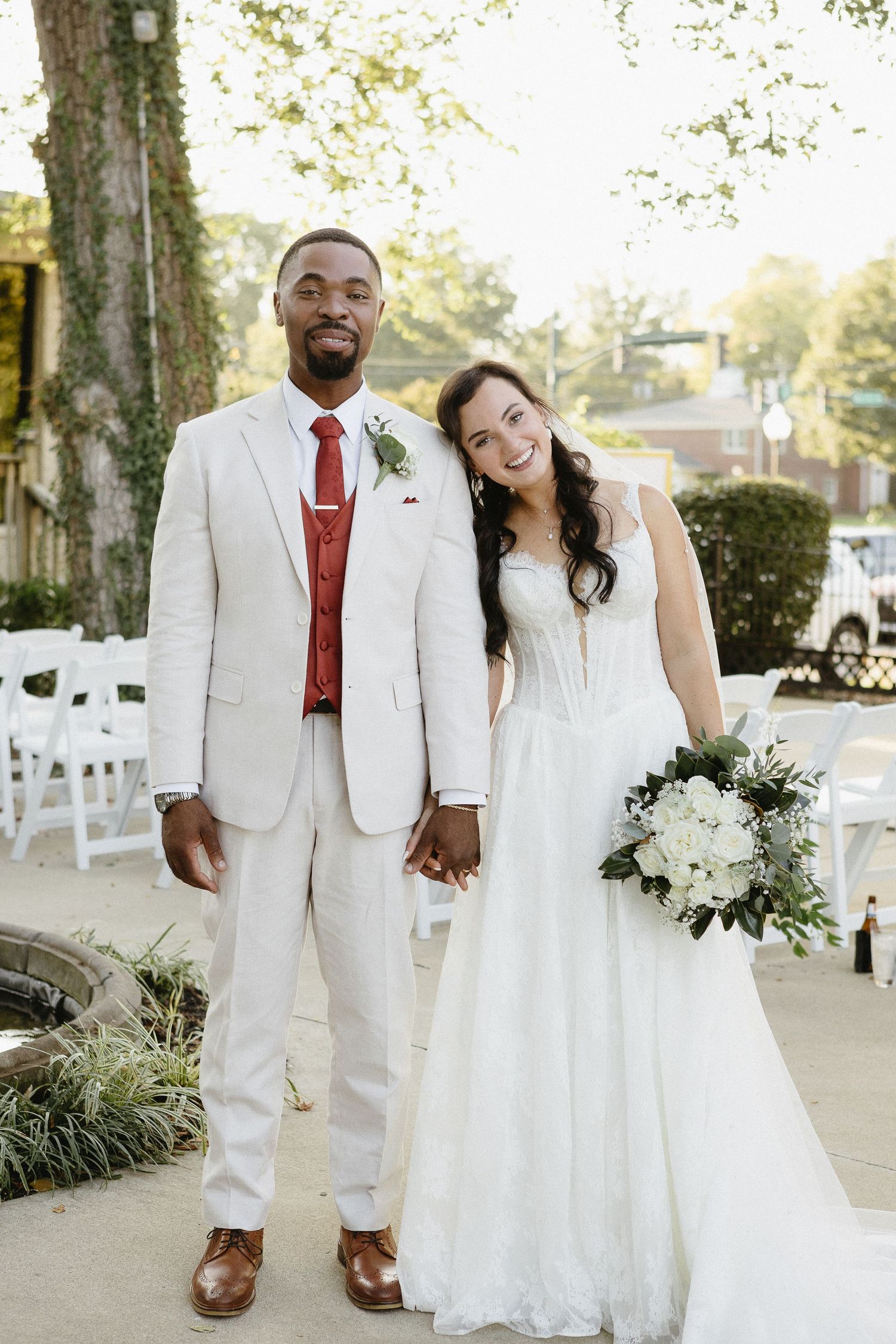 A wedding couple in coordinating white attire pose together at their outdoor venue.