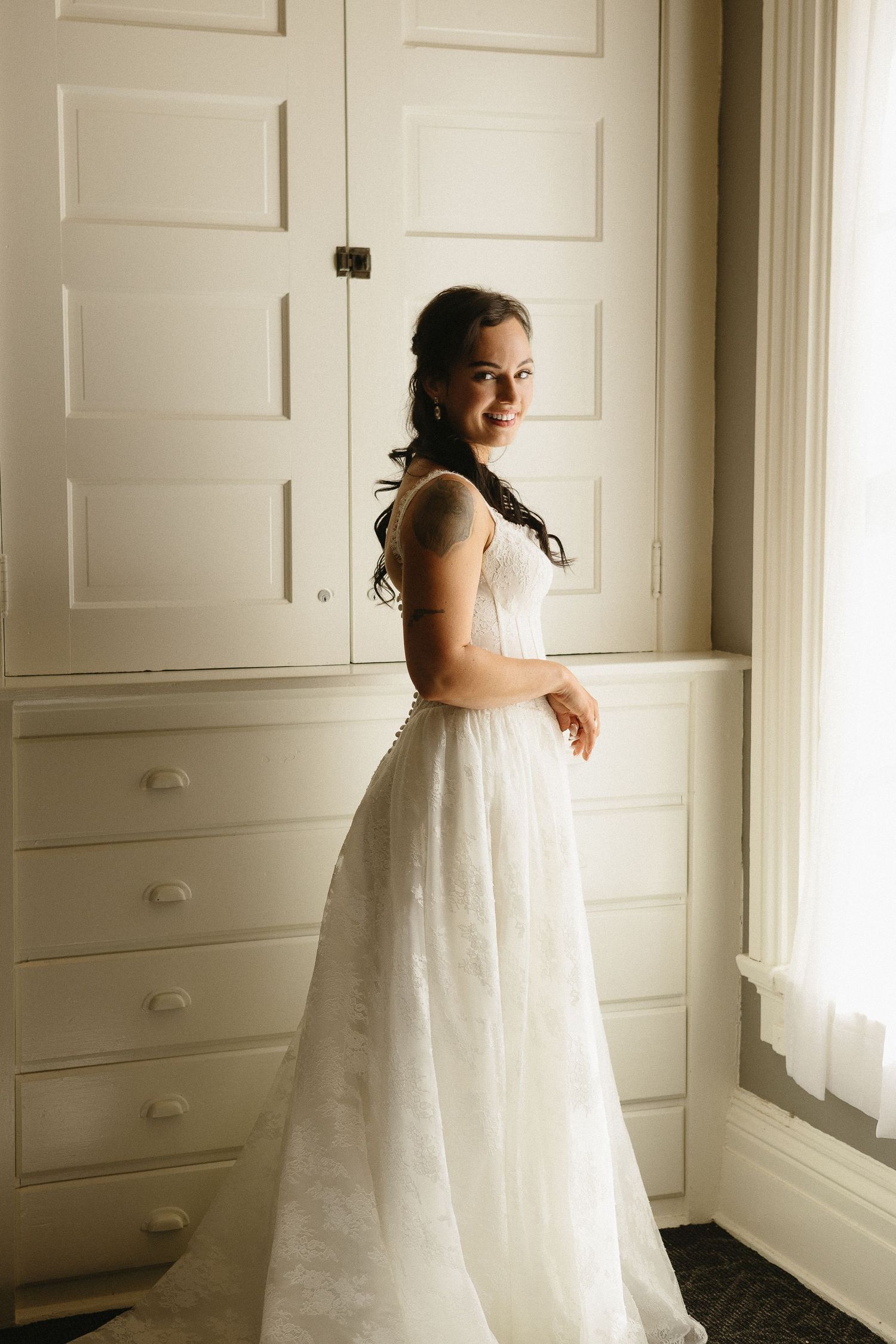 A bride in a flowing white wedding gown stands by a window in a bright, minimalist room with white walls and doors.