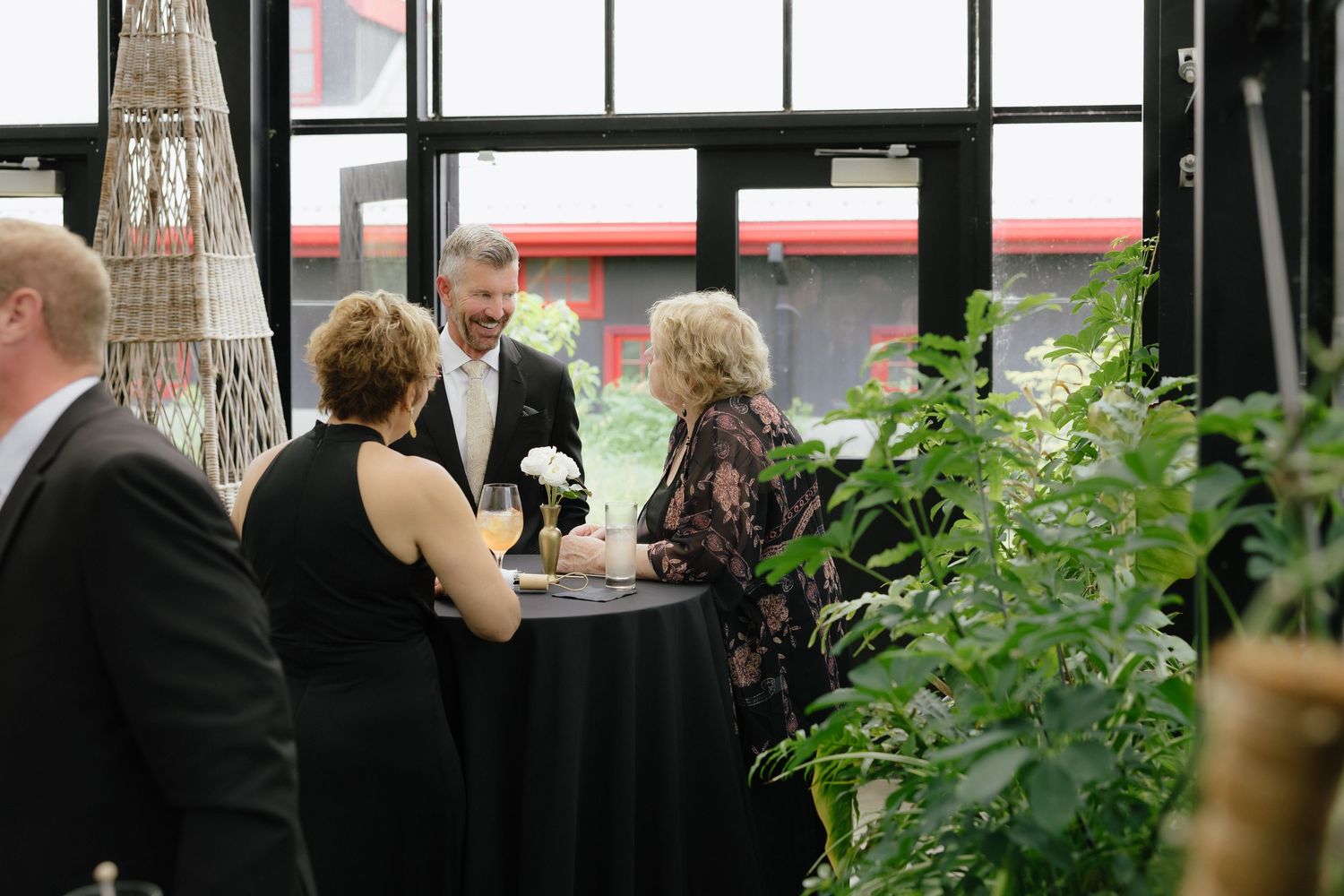 Guests in formal attire mingle near lush green plants at an elegant indoor wedding reception.