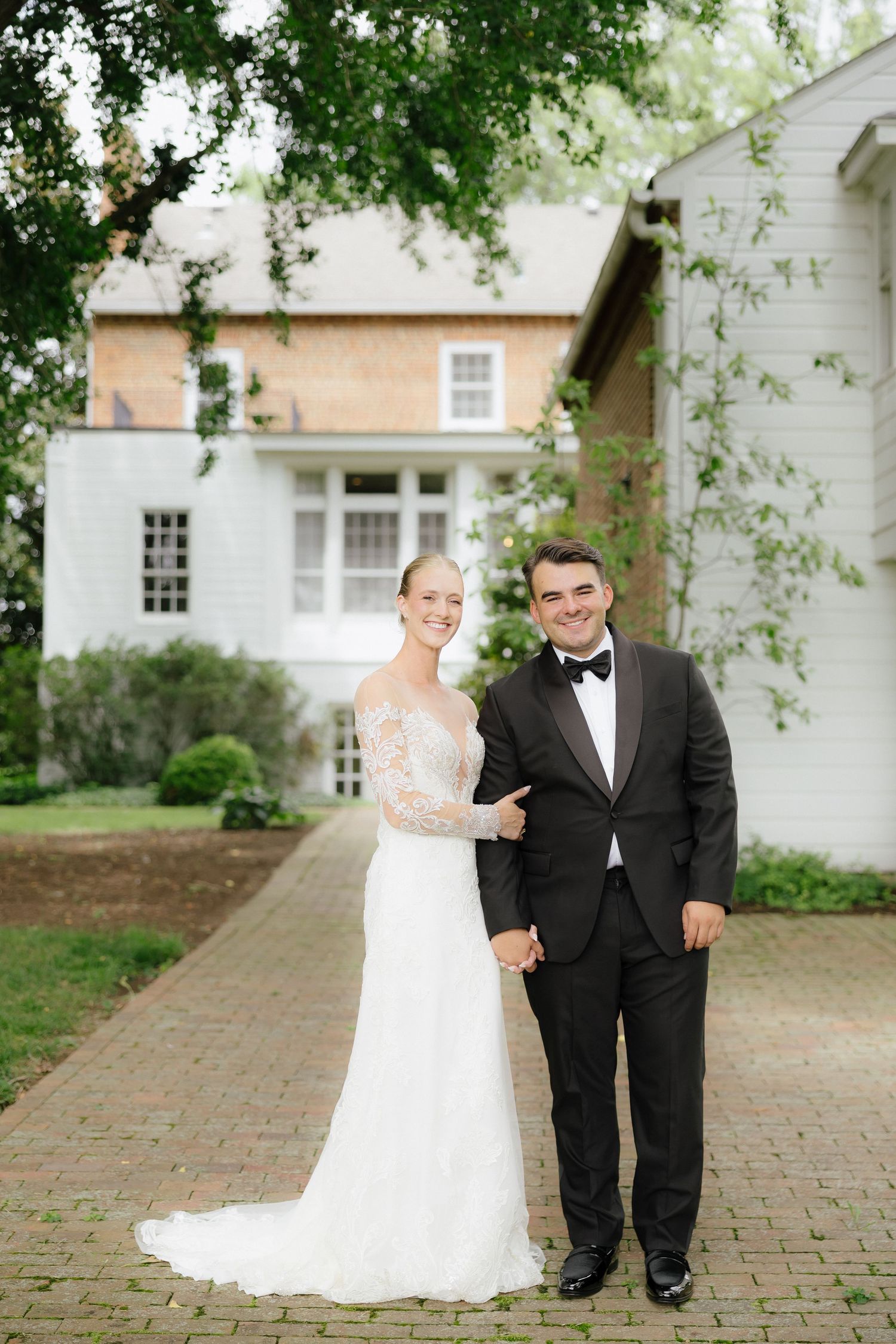 Bride in lace long-sleeve wedding gown and groom in black tuxedo pose together outside a white colonial home.