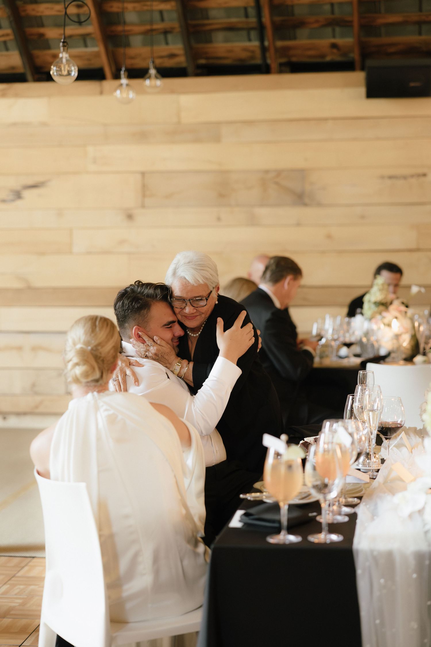 Groom embracing an older woman at a wedding reception table set with champagne glasses in a rustic barn venue.