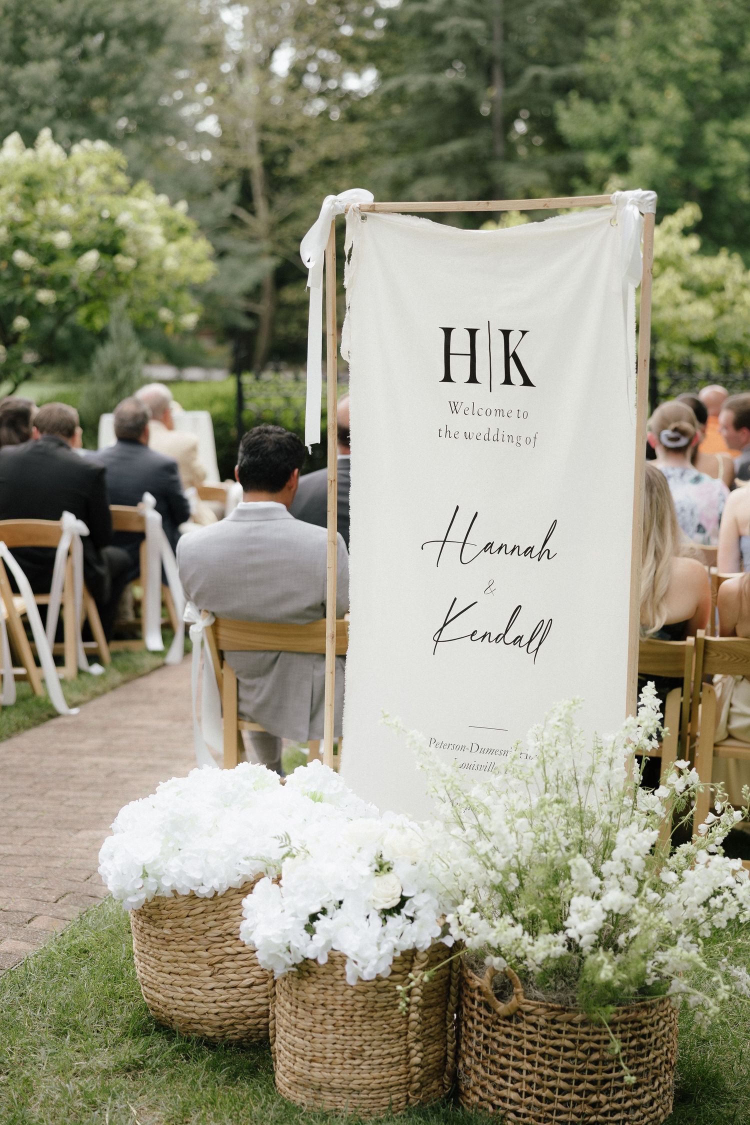 Outdoor wedding ceremony welcome banner for Hannah and Kendall with white floral arrangements in baskets.