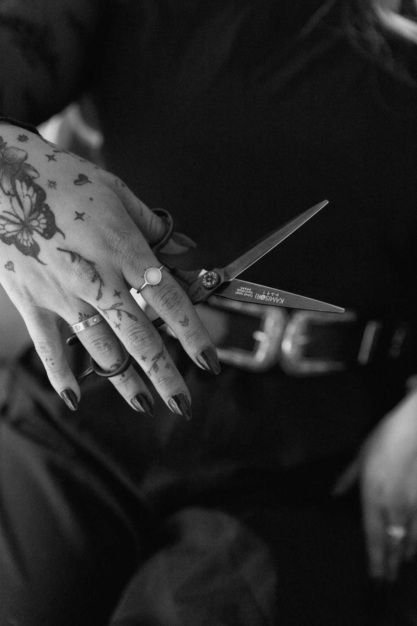 Tattooed hands with dark nails holding scissors, rings visible, black and white photo of a hairstylist.