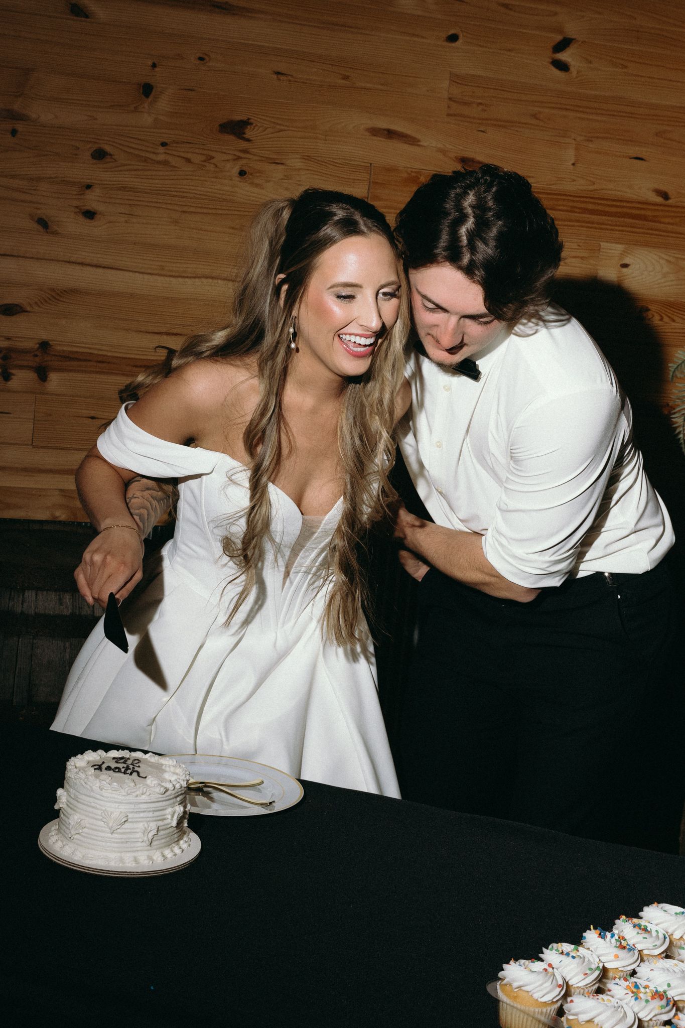 Wedding party shares casual moments and laughter at wooden table with cupcakes.