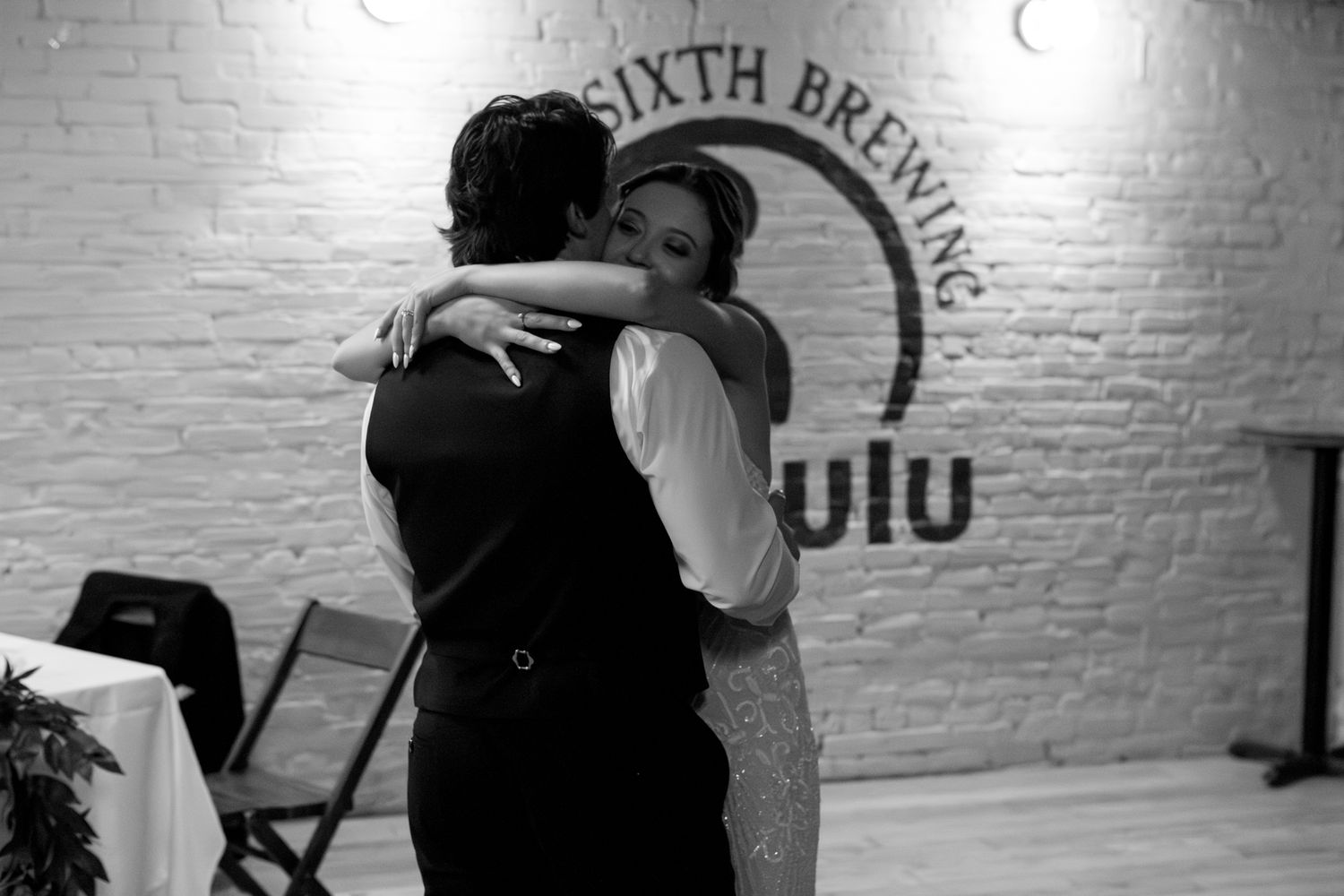 A black and white photograph of a couple sharing an intimate dance at a wedding reception by a brick wall.