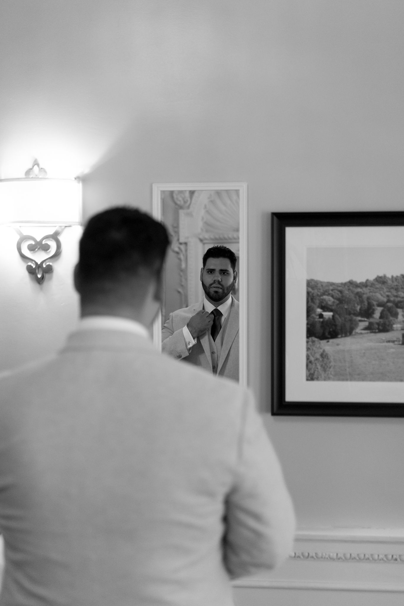 Black and white image showing a reflection in a mirror with a framed landscape photograph on the wall.