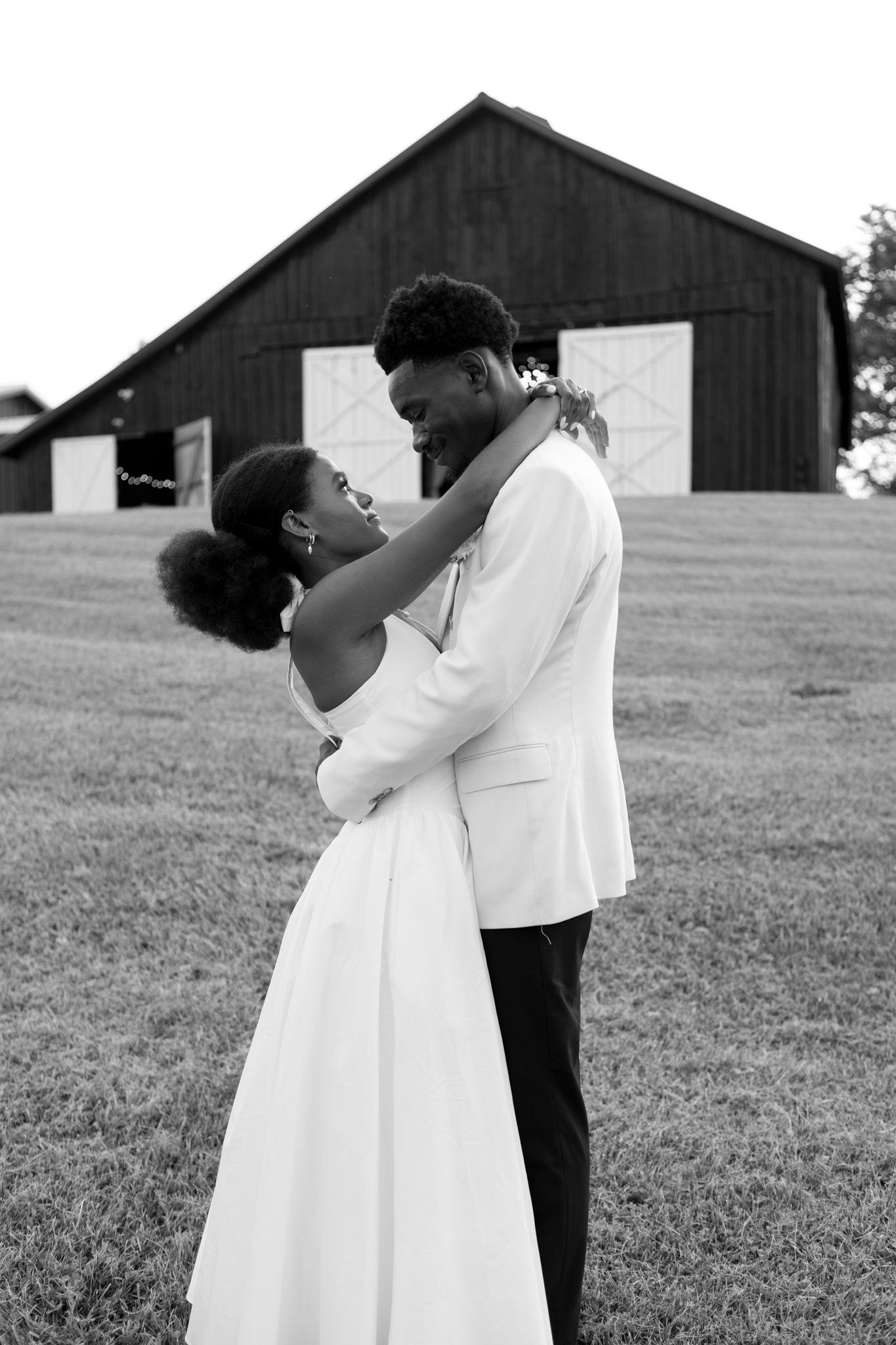 A romantic black and white portrait of newlyweds embracing in front of a rustic barn.