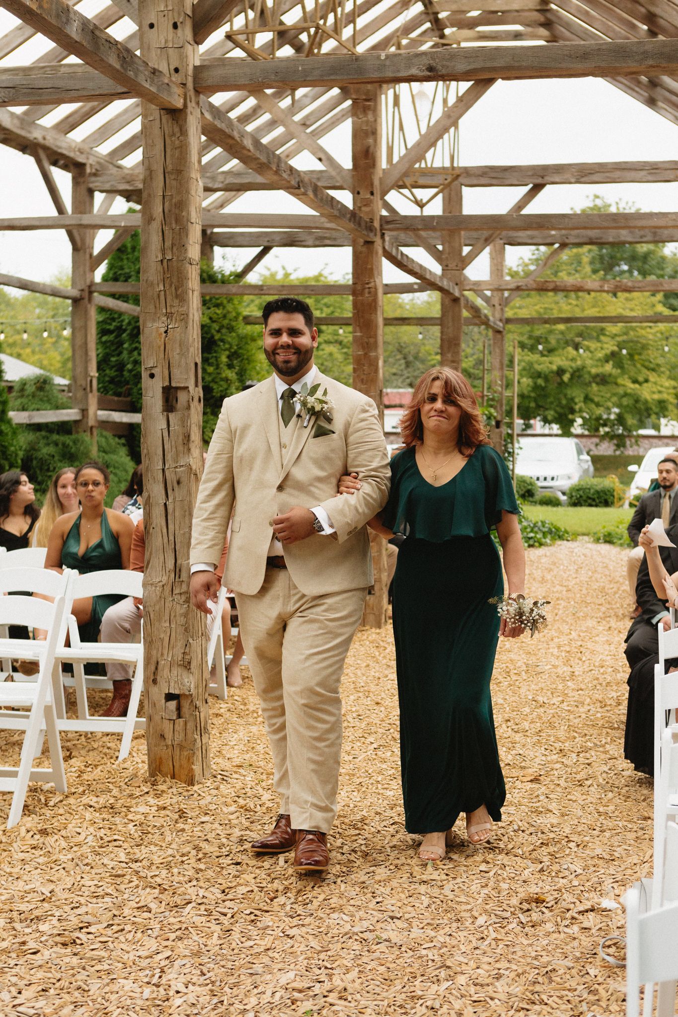Wedding attendees walk down a mulch-covered aisle under a rustic wooden beam structure at an outdoor venue.