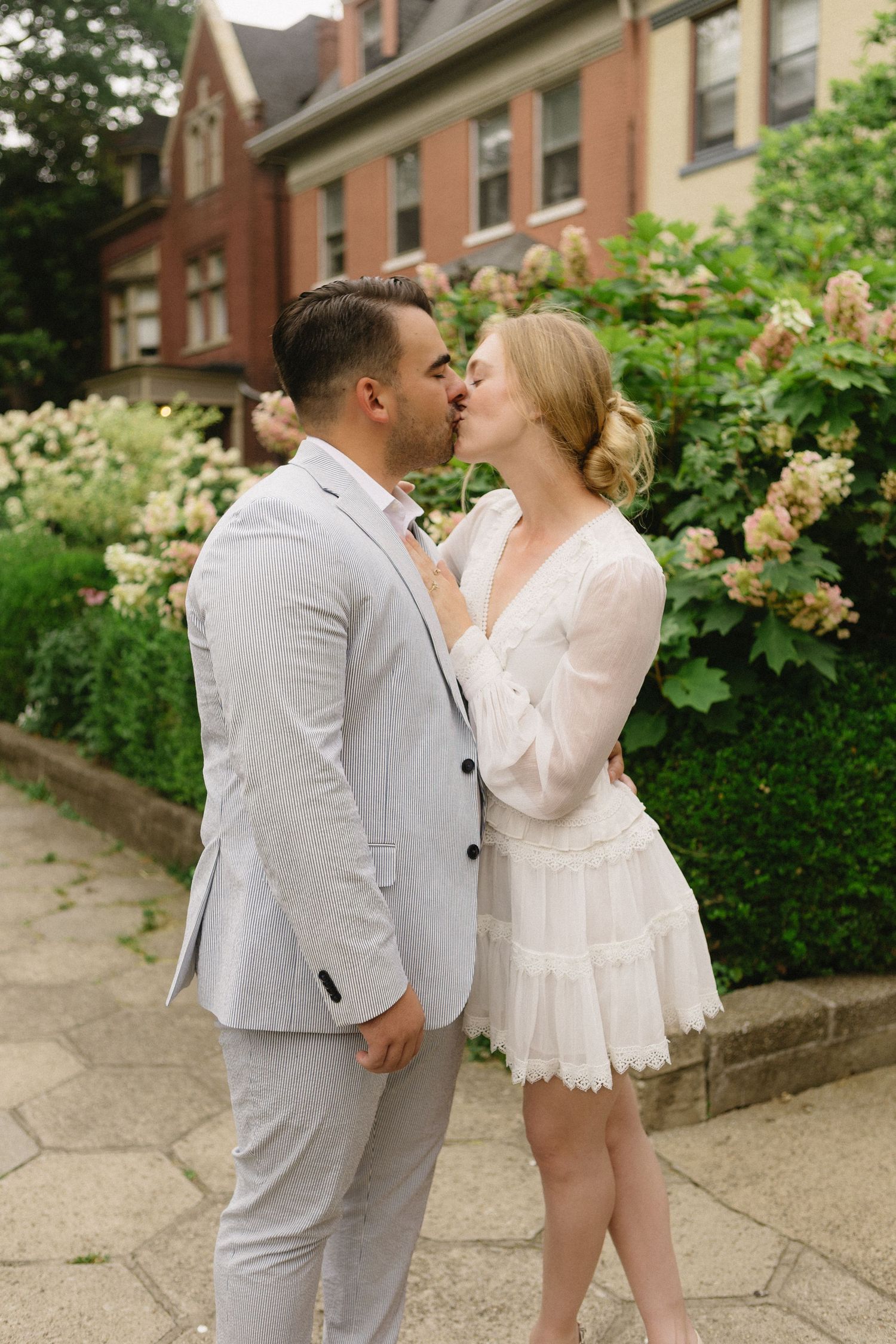 Couple sharing a romantic kiss outdoors in wedding attire, surrounded by lush greenery and a brick building backdrop.