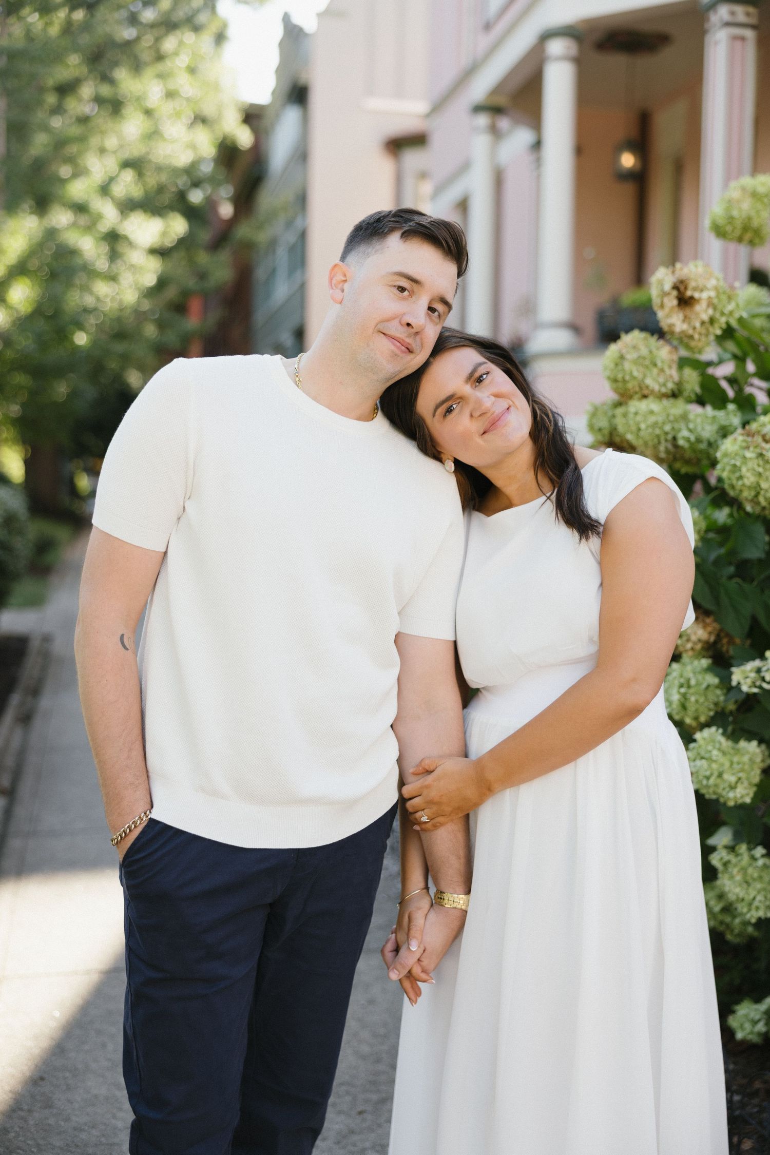 Couple in matching cream outfits embrace tenderly beside blooming hydrangeas in an urban setting.