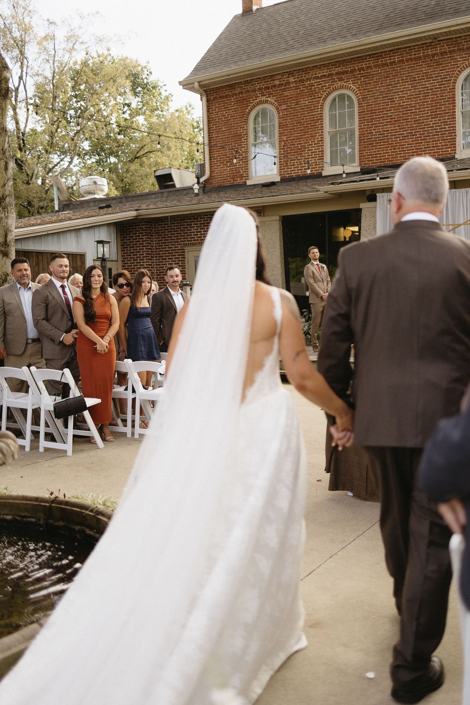 Wedding ceremony setup outside brick building with white chairs and gathered guests.