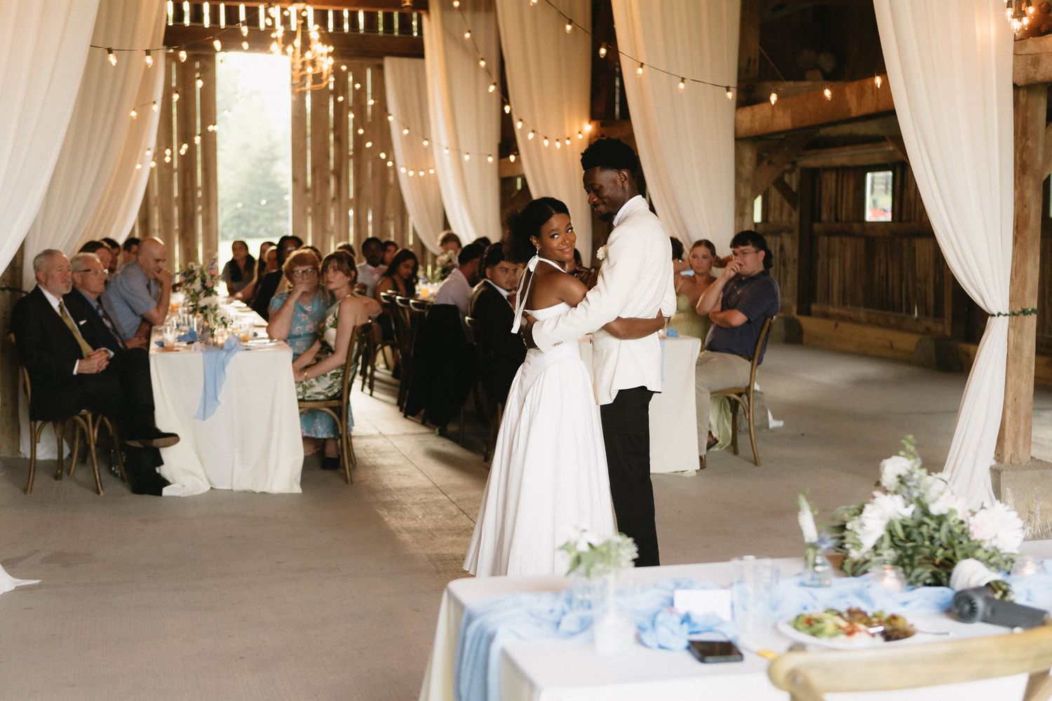 Wedding reception venue decorated with elegant white drapes and columns filled with guests seated at round tables.