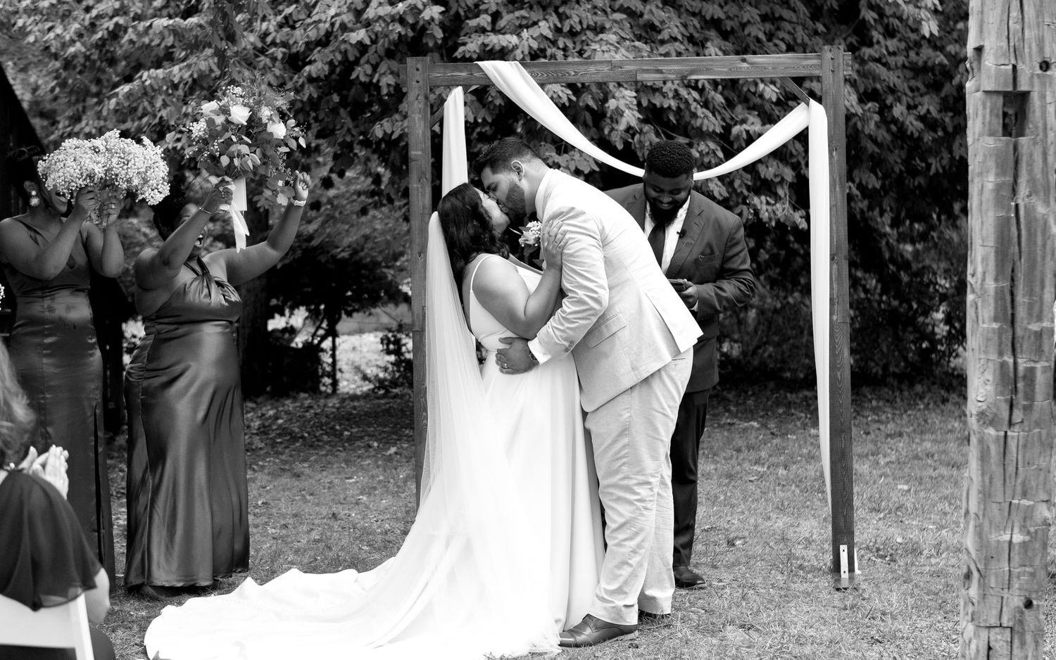 A romantic black and white outdoor wedding ceremony under a simple wooden arch decorated with white fabric.