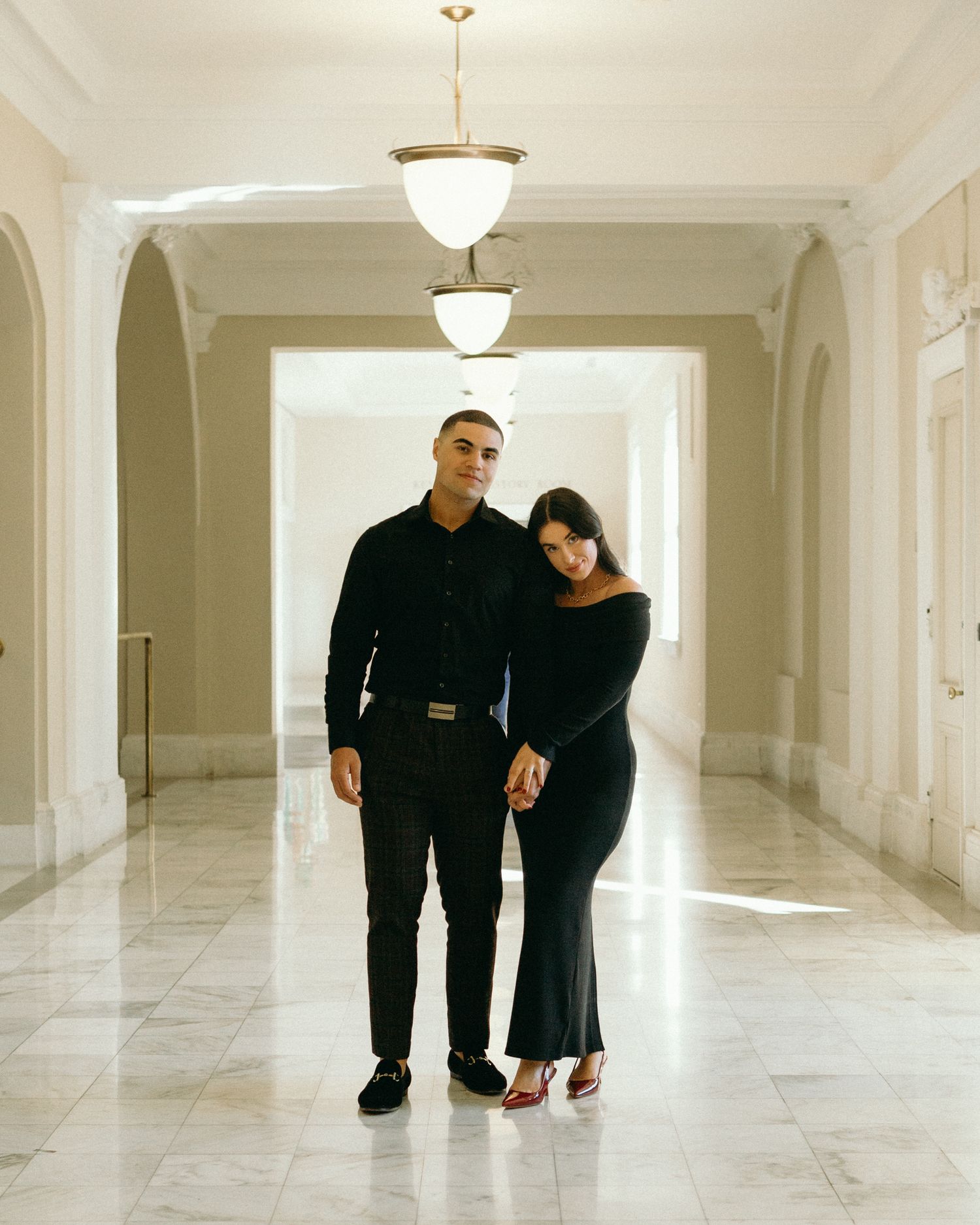 Stylish couple in all black outfits pose together in an elegant marble hallway with arches and pendant lights.