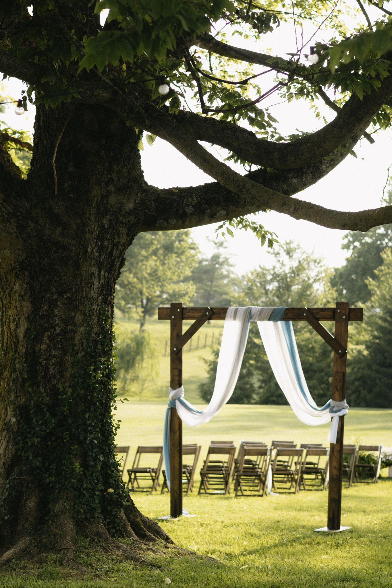 Outdoor wedding ceremony setup with white curtains draped over wooden posts under large trees in a serene garden setting.