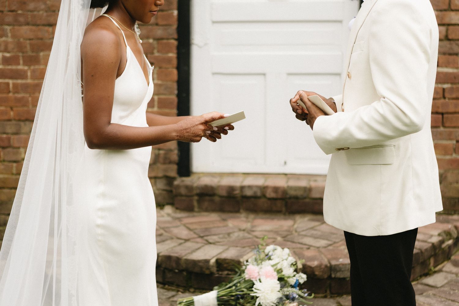 Elegant couple in wedding attire stands against white door and brick wall.
