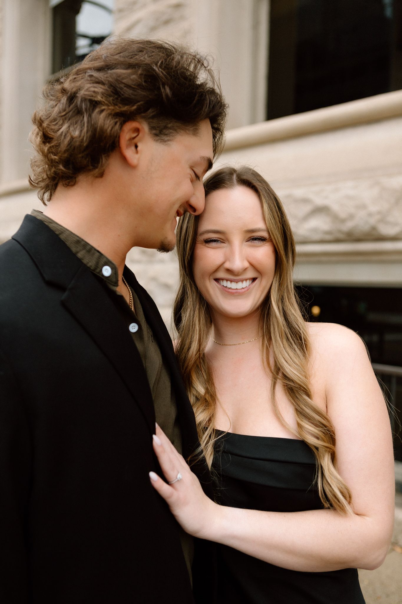 Engaged couple smiling and embracing, with the woman showing her engagement ring, captured in downtown Louisville