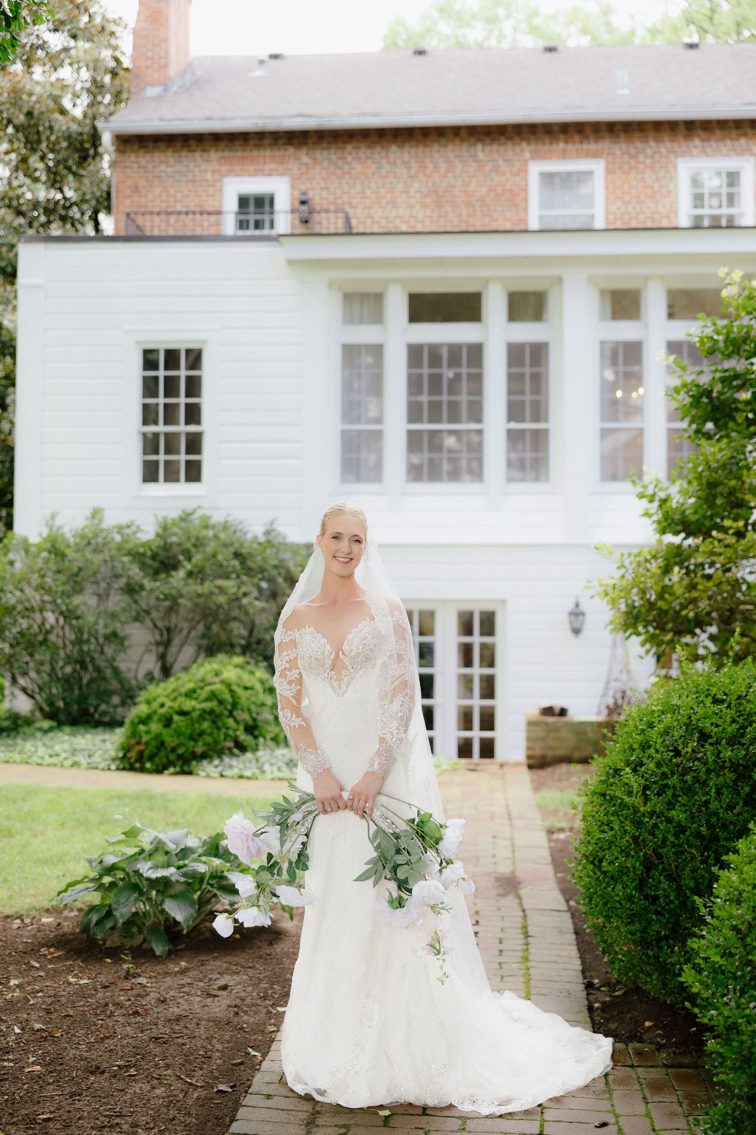 Elegant bride in lace long-sleeve wedding gown holding greenery bouquet standing before historic brick manor house.