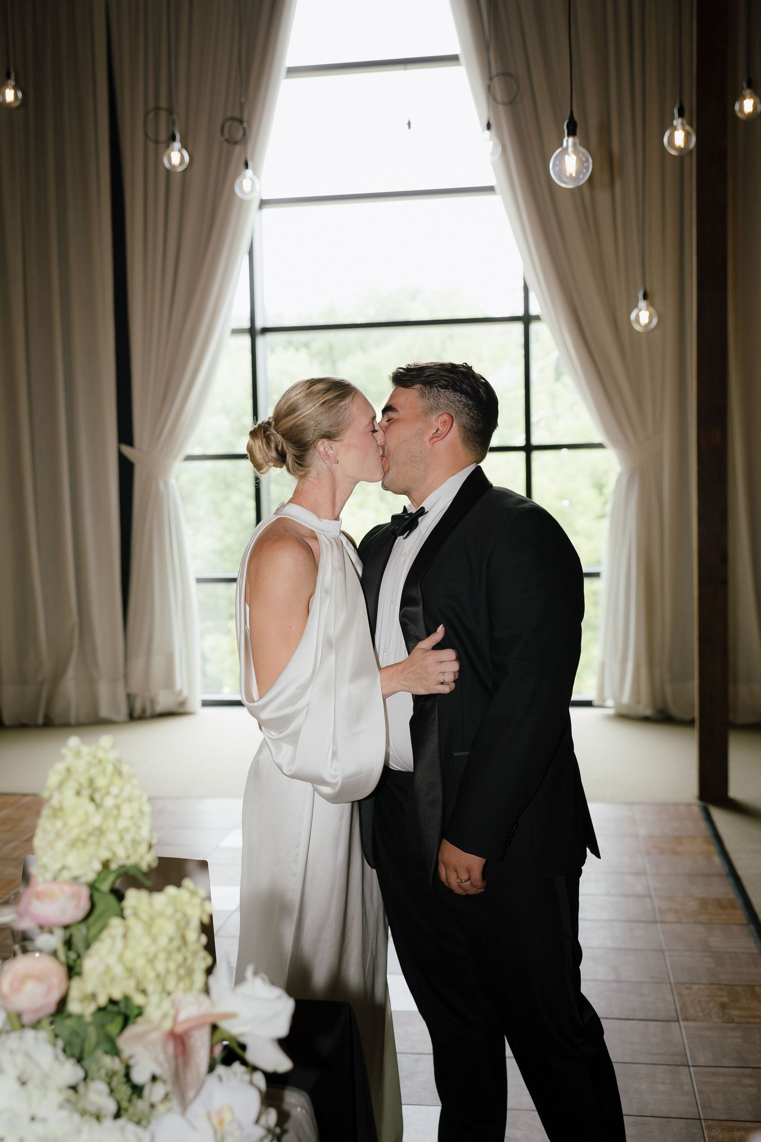 Bride and groom share first kiss at elegant wedding ceremony with draped curtains and Edison bulb lighting.