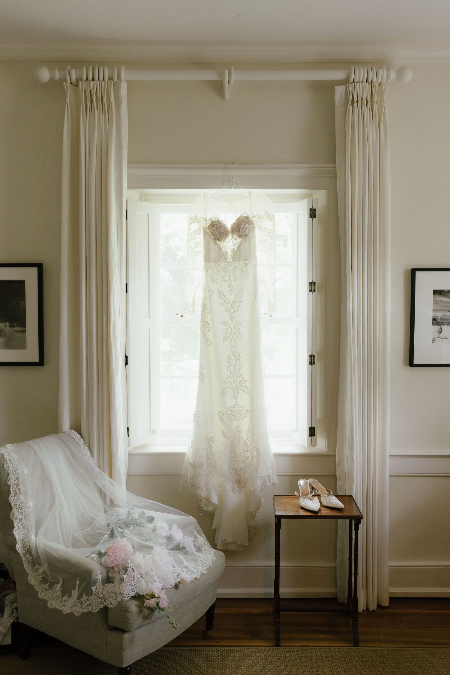 Elegant lace wedding dress hanging in sunlit window with bridal accessories, white heels, and floral bouquet on chair nearby.